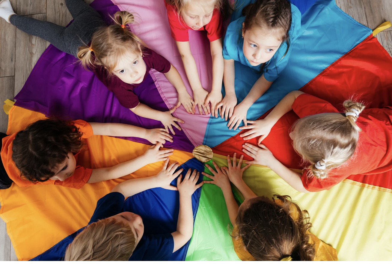 Children sitting in a circle on a multicolored parachute, touching the center with their hands.