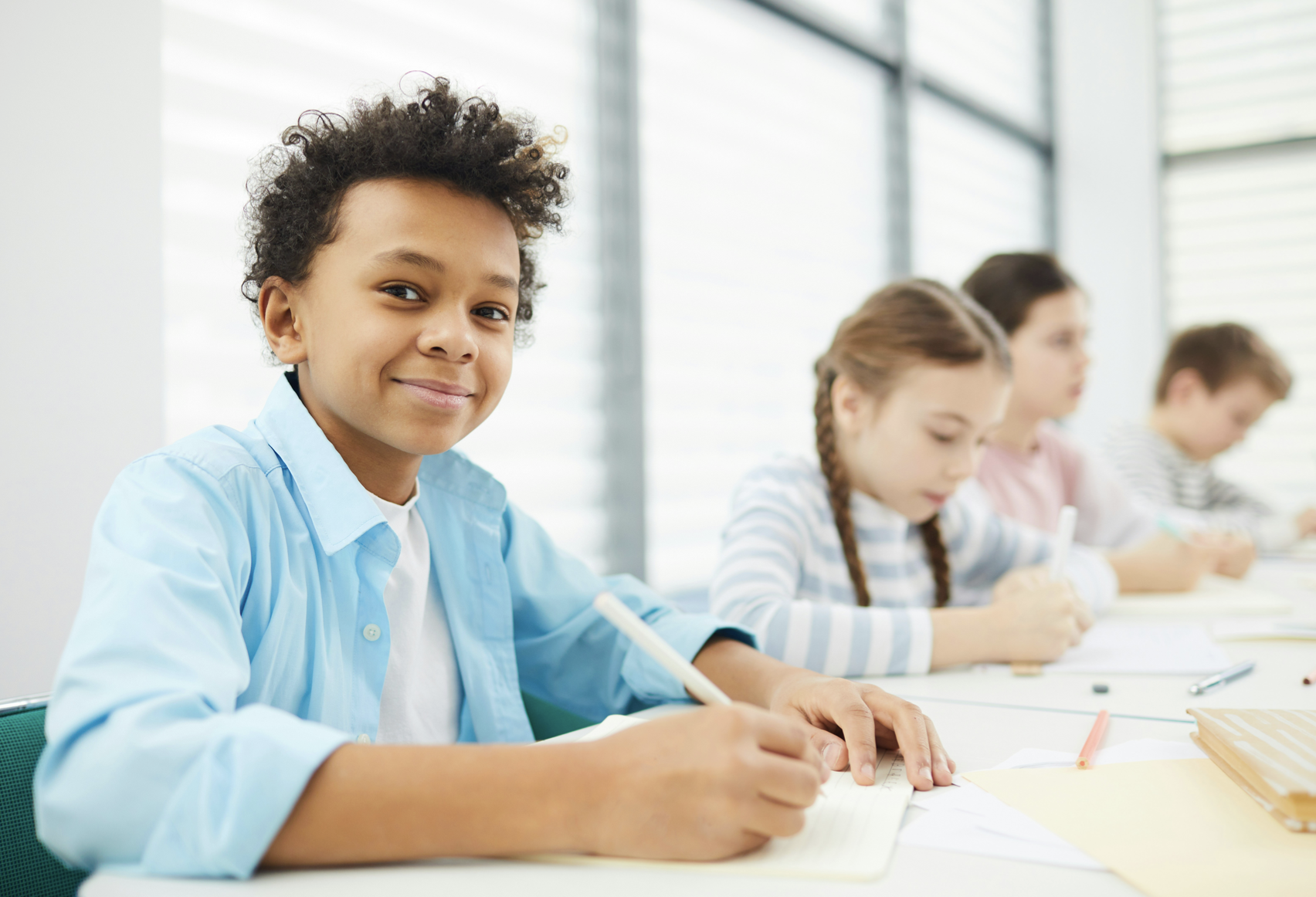 Young boy with curly hair smiling while sitting in a classroom with other children, writing on paper.