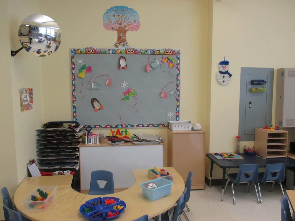 A classroom with a light yellow wall, decorated with a colorful tree and an art display with penguin and snowflake decorations. Blue tables and chairs are arranged for children, with craft supplies and art projects on the surfaces. A star-shaped mirr