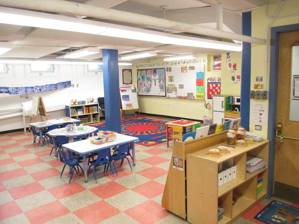 A classroom with small tables and chairs, a colorful rug, shelves with books and toys, and a bulletin board with children's artwork.