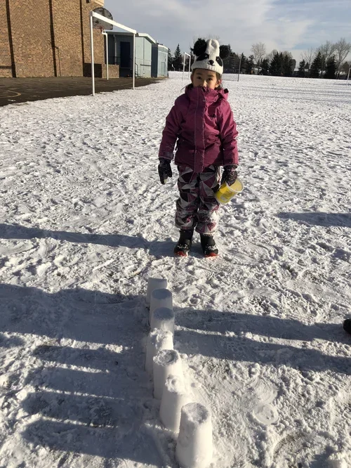 A young girl in a purple winter jacket, snow hat with panda face, and patterned snow pants standing on snow-covered ground at a playground, holding a yellow cup, with a snow sculpture in front of her.