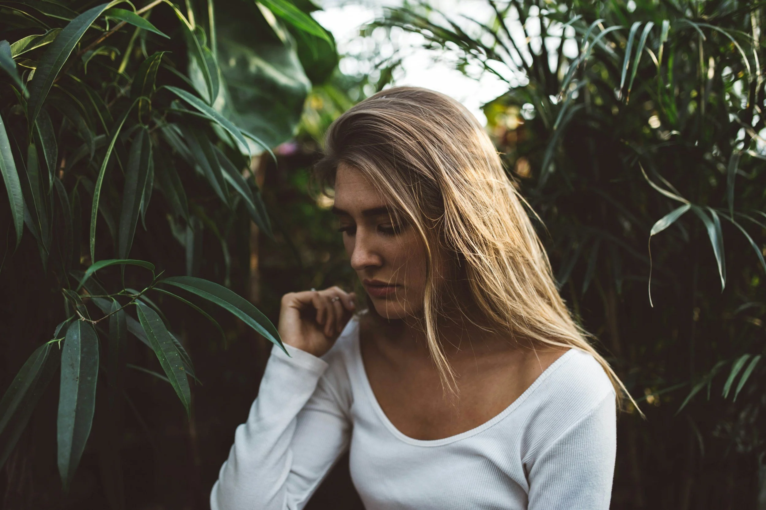 A woman with long blonde hair and a white shirt standing among tall, green plants in a lush, outdoor setting.
