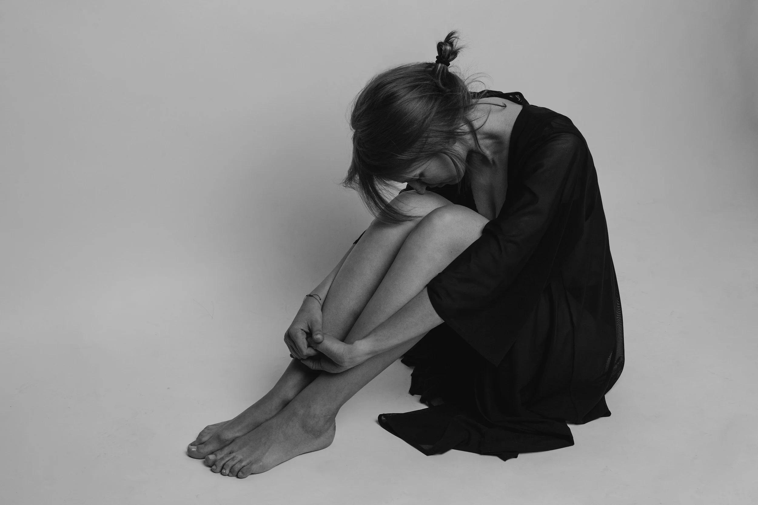 A woman with short hair sitting on the floor, wearing a black dress, holding her knees to her chest with her head bowed down and her face hidden.