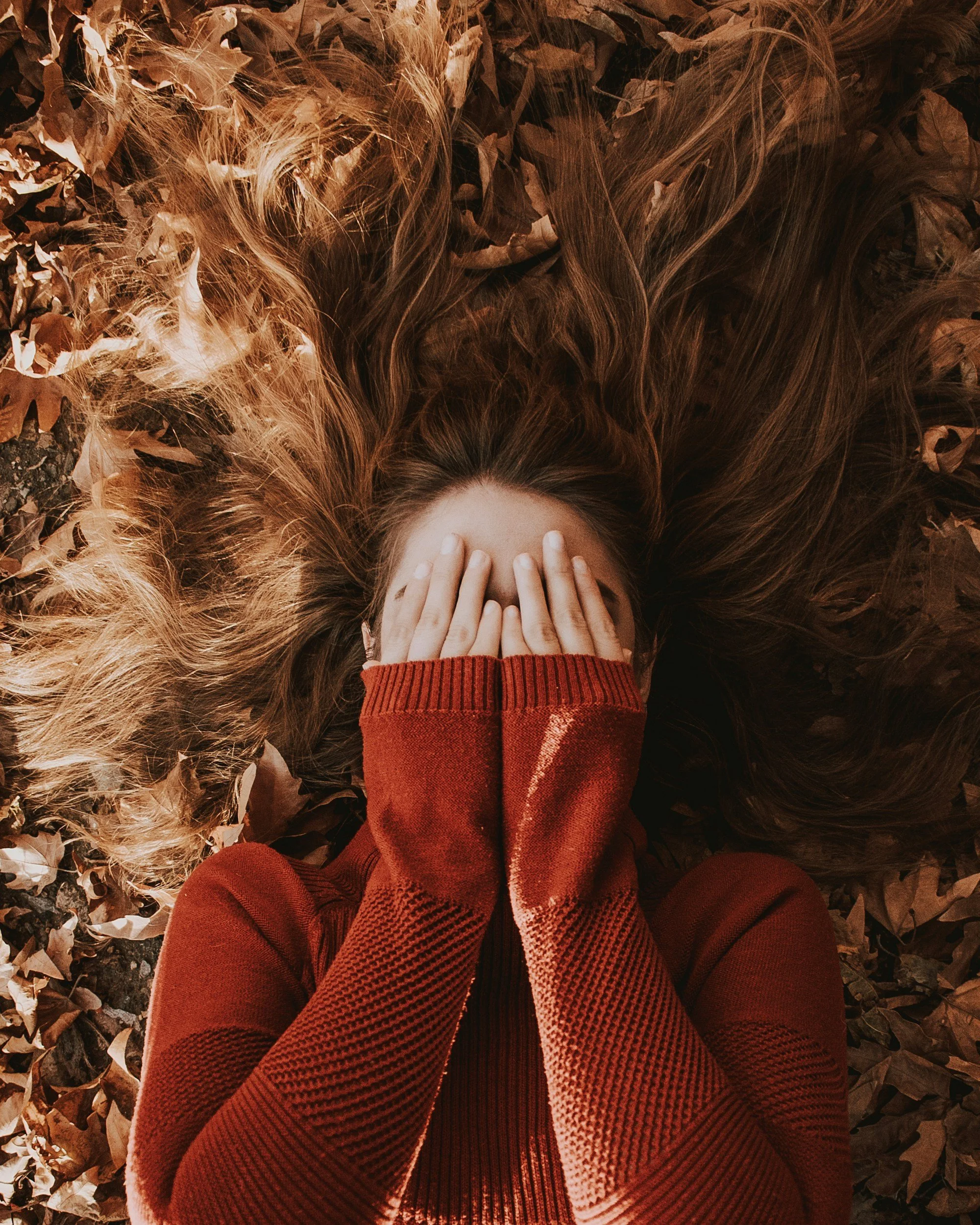 A woman with long hair lying on the ground covered with brown fallen leaves, covering her face with her hands, wearing a red sweater.