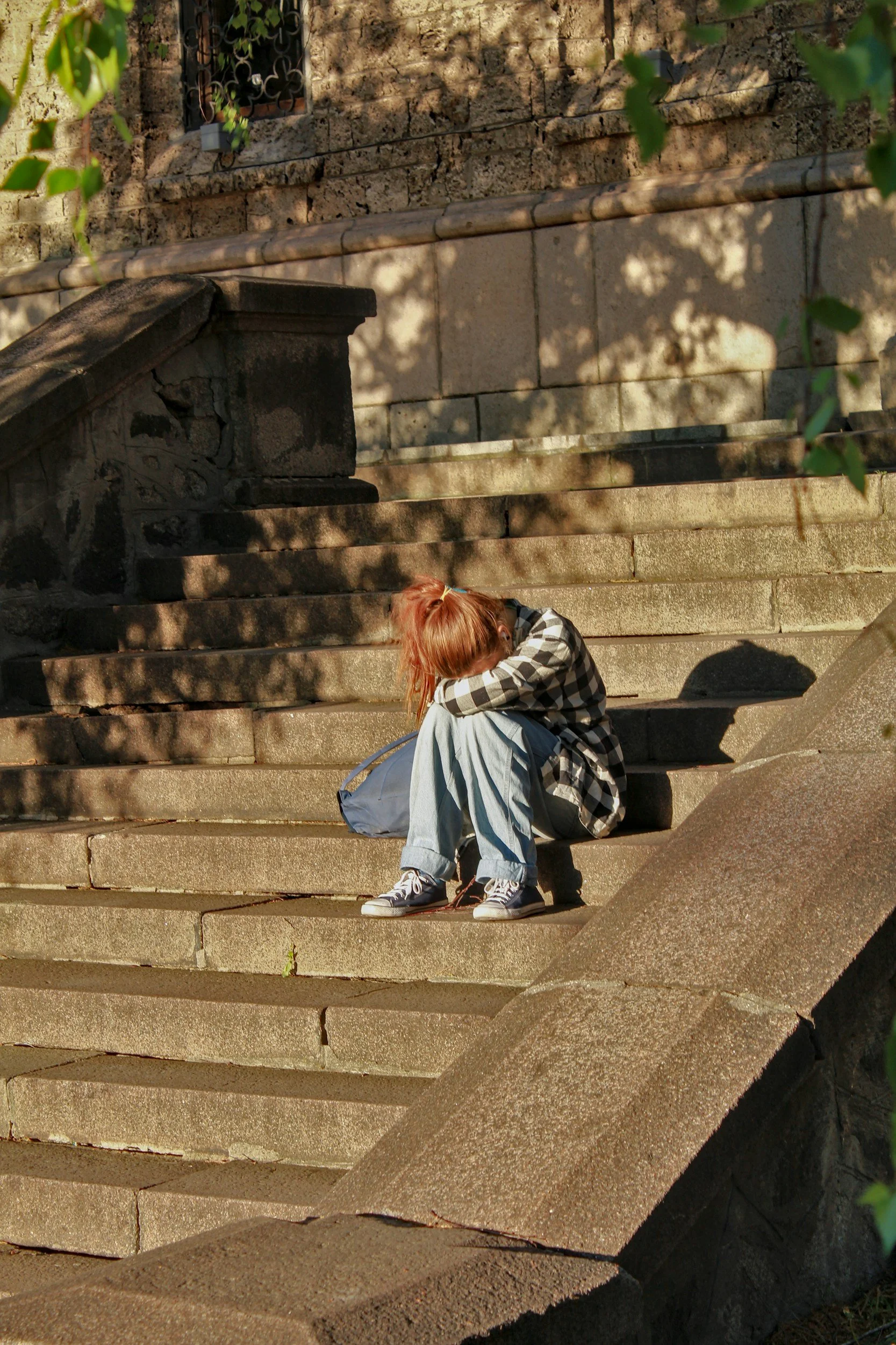 A person with red hair sitting on stone stairs, resting their head on their knees, and carrying a blue bag, surrounded by sunlight and shadows.