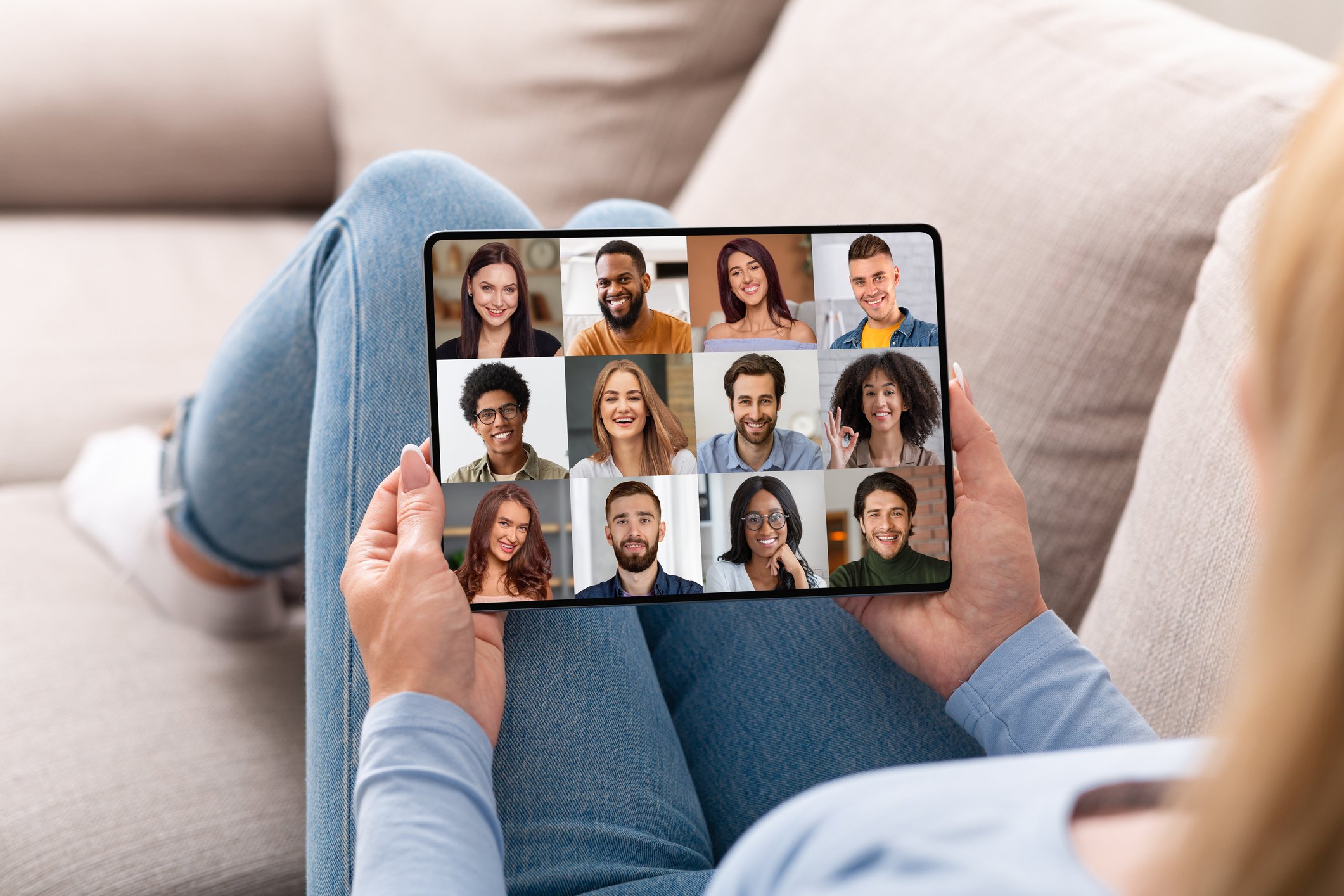 Person sitting on a beige sofa holding a tablet with a video call of multiple diverse smiling people.