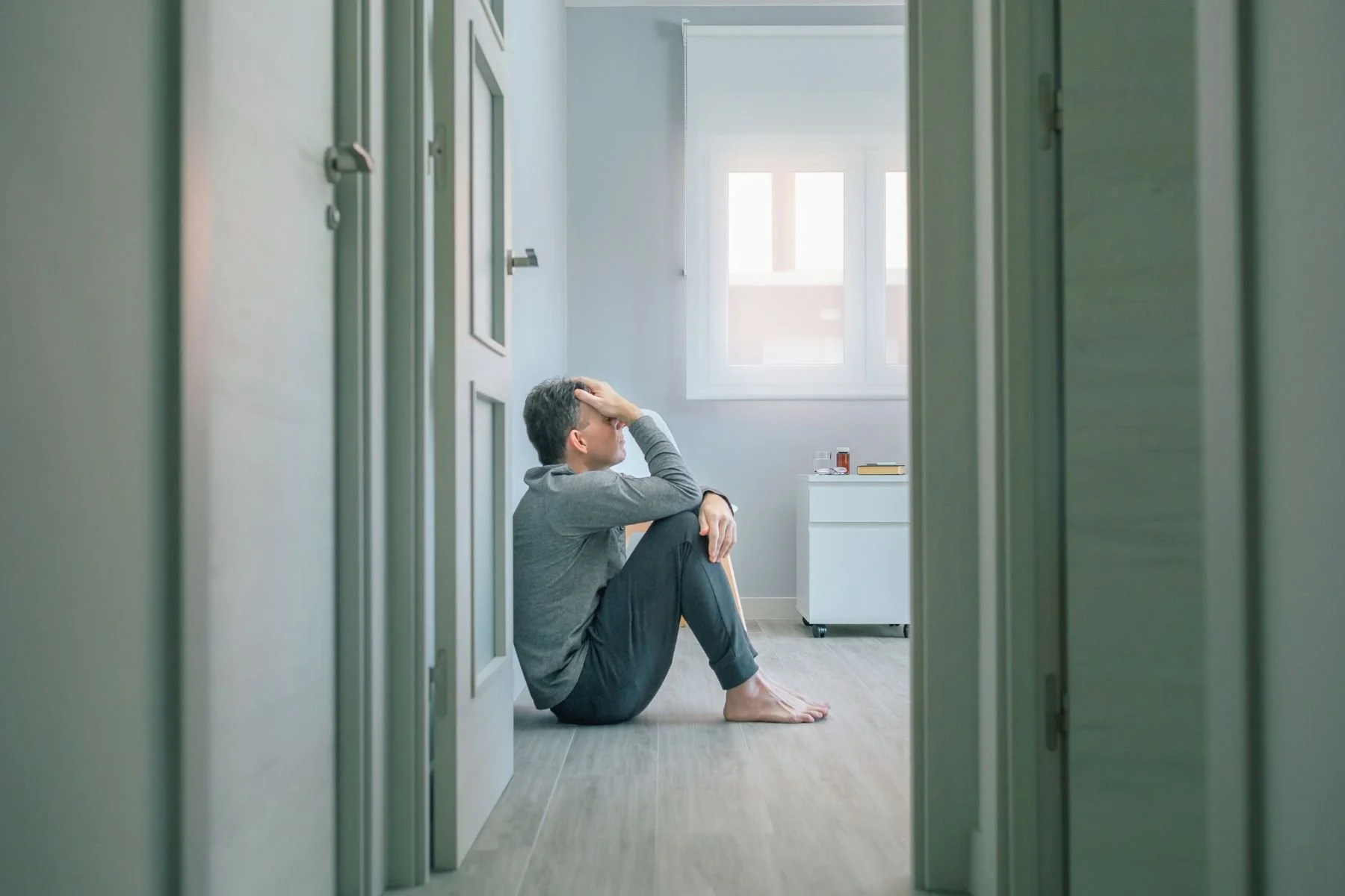 A man sitting on the floor of a room, holding his head in distress, seen through a doorway with open doors.