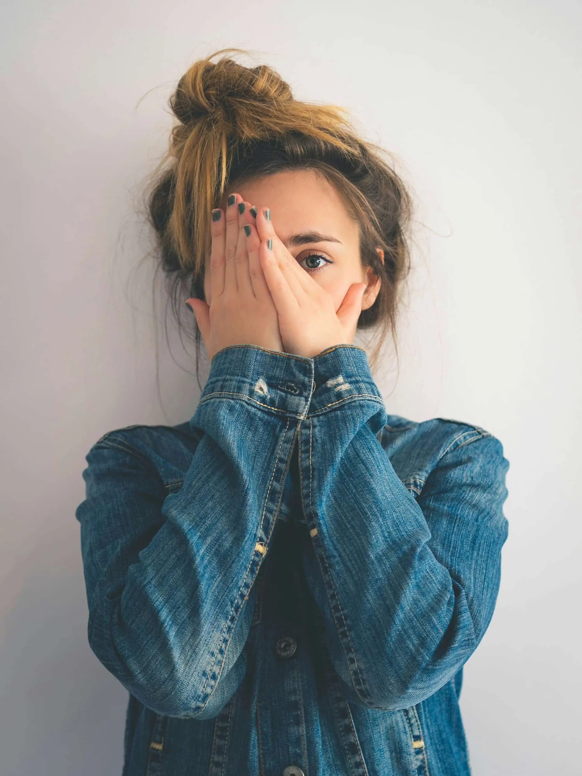 A woman with light brown hair tied in a messy bun, covering part of her face with her hand, wearing a distressed denim jacket, against a plain white background.