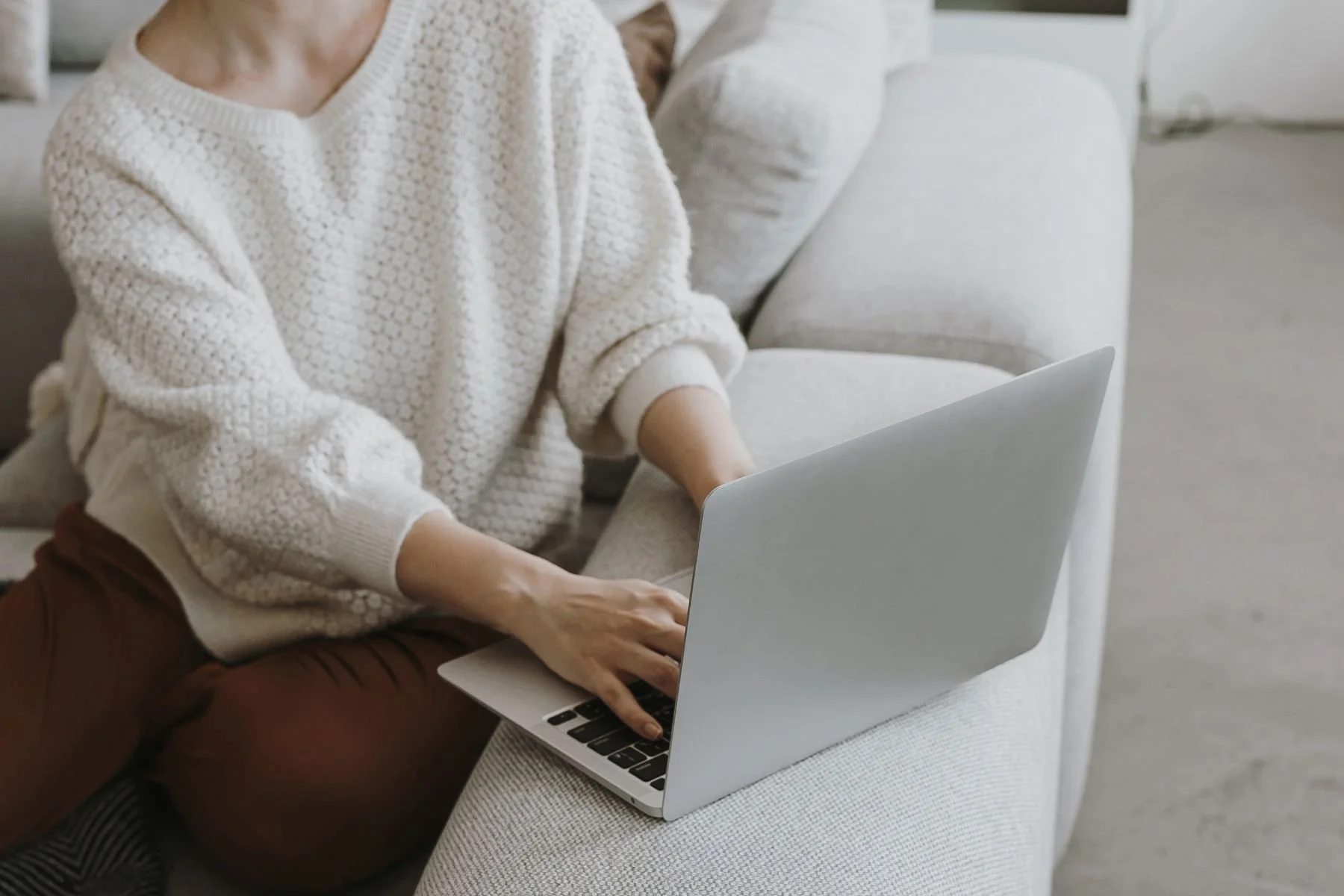 Person wearing a cream-colored textured sweater working on a silver laptop on a light gray sofa.
