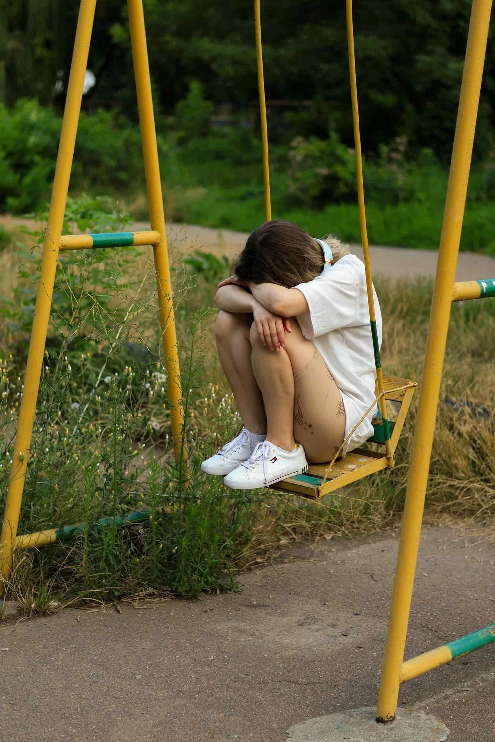 Child sitting on a yellow playground swing, hugging her knees and resting her head on her arms, in a grassy outdoor area.