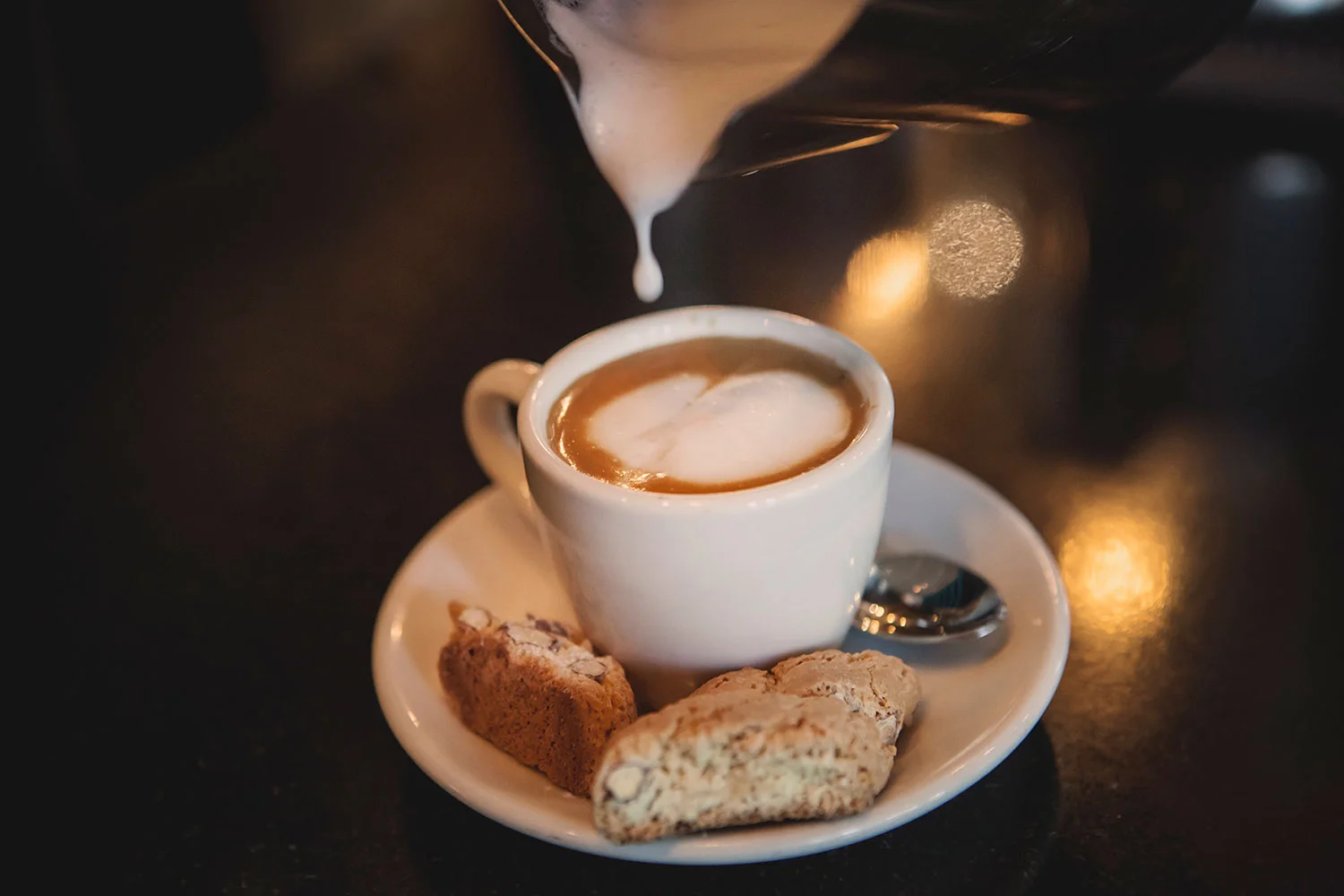 A cup of cappuccino with foam art on top, placed on a saucer with a small spoon and two cookies, one chocolate chip and one biscotti, on a dark table.