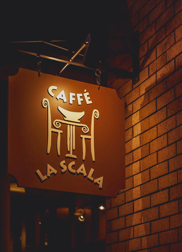 Sign for Caffé La Scala mounted on a brick wall, featuring a stylized logo with a bowl on top of columns and decorative scrolls.