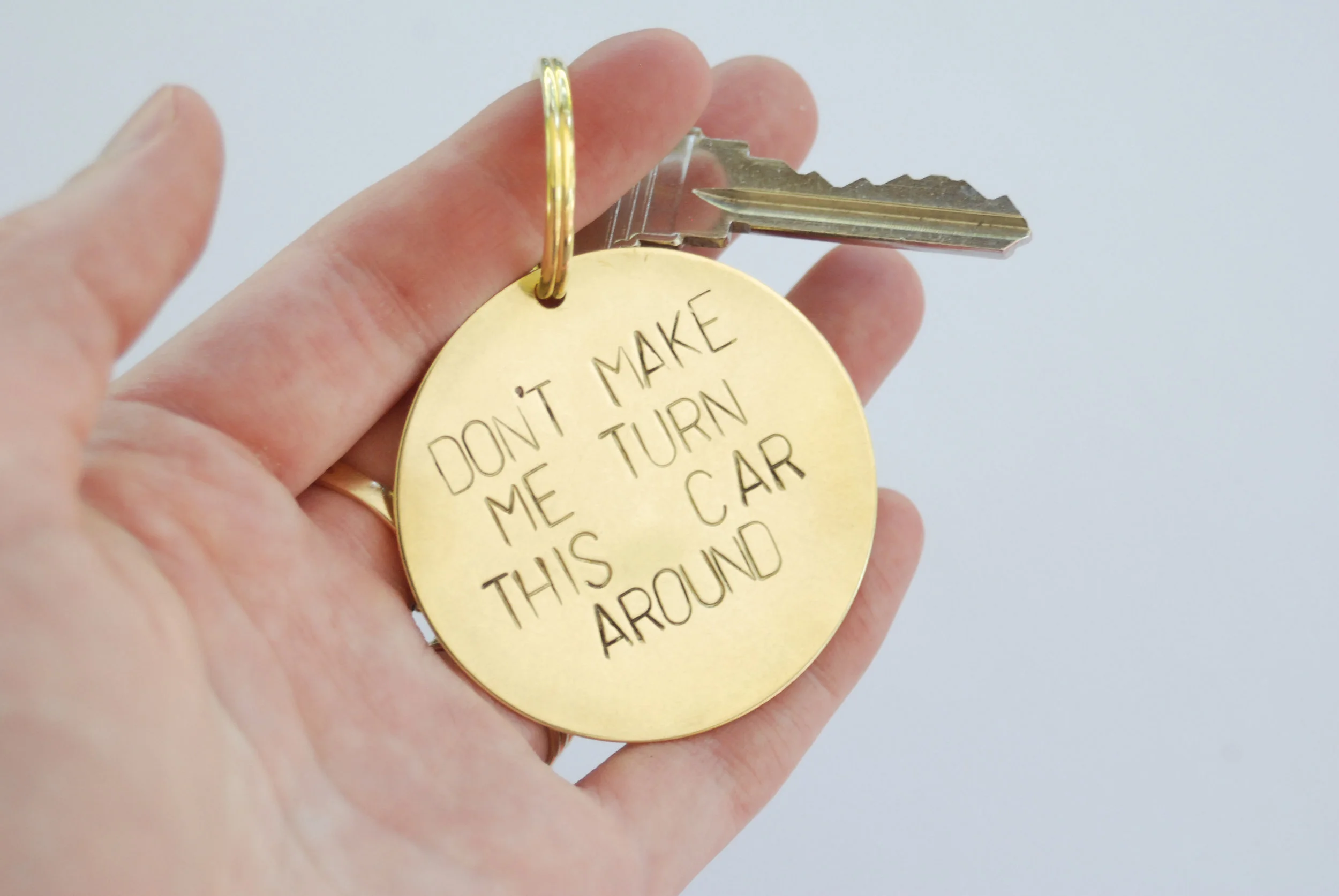 A brass circle keychain stamped with the phrase "don't make me turn this car around"