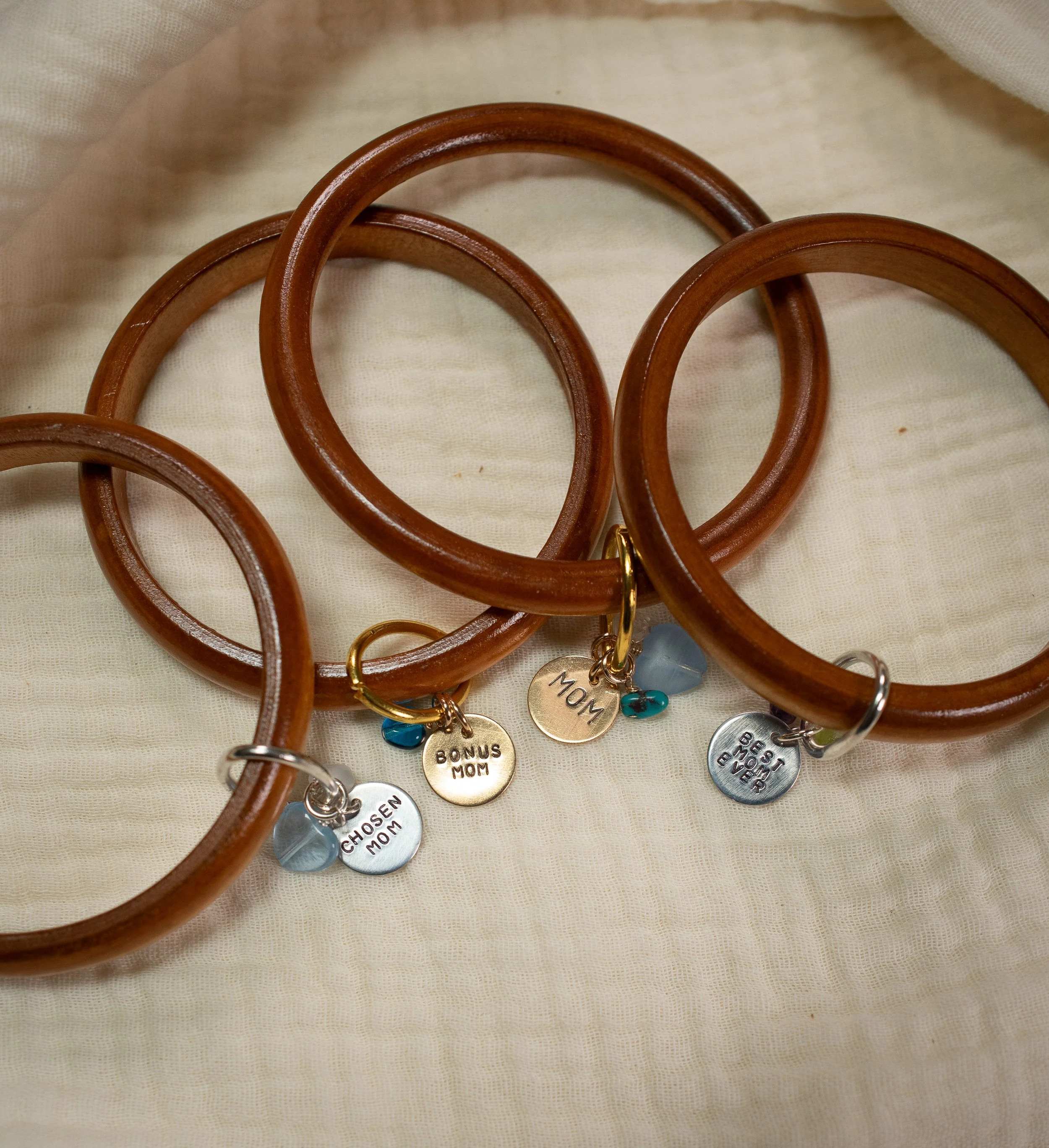 Four wooden bangles are stacked over each other and have charms that say "chosen mom," "bonus mom", "mom"
