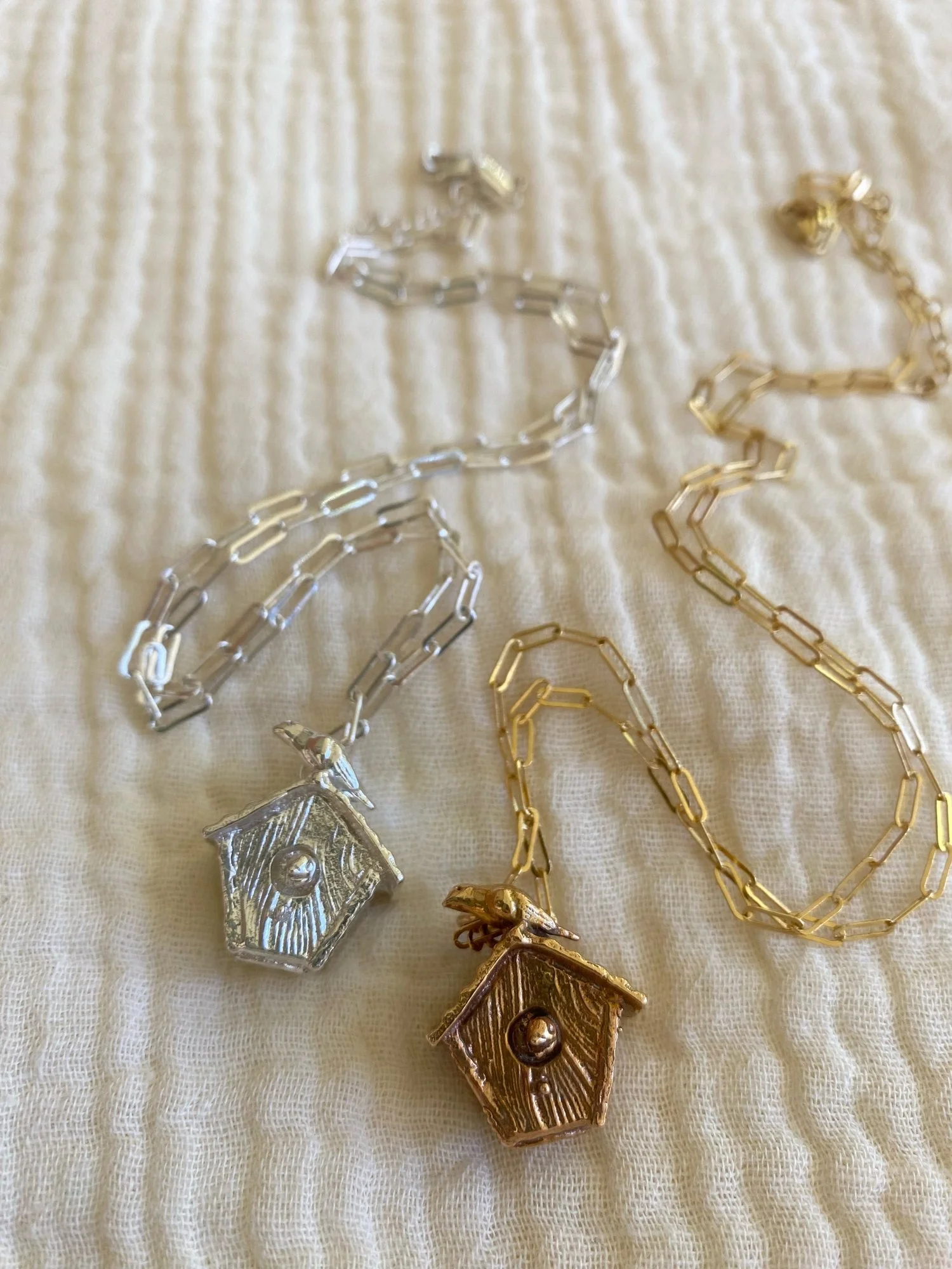 A bronze and a silver necklace on a white background: they have pendants shaped like little bird houses with a mother bird on top and a baby bird poking its head out.