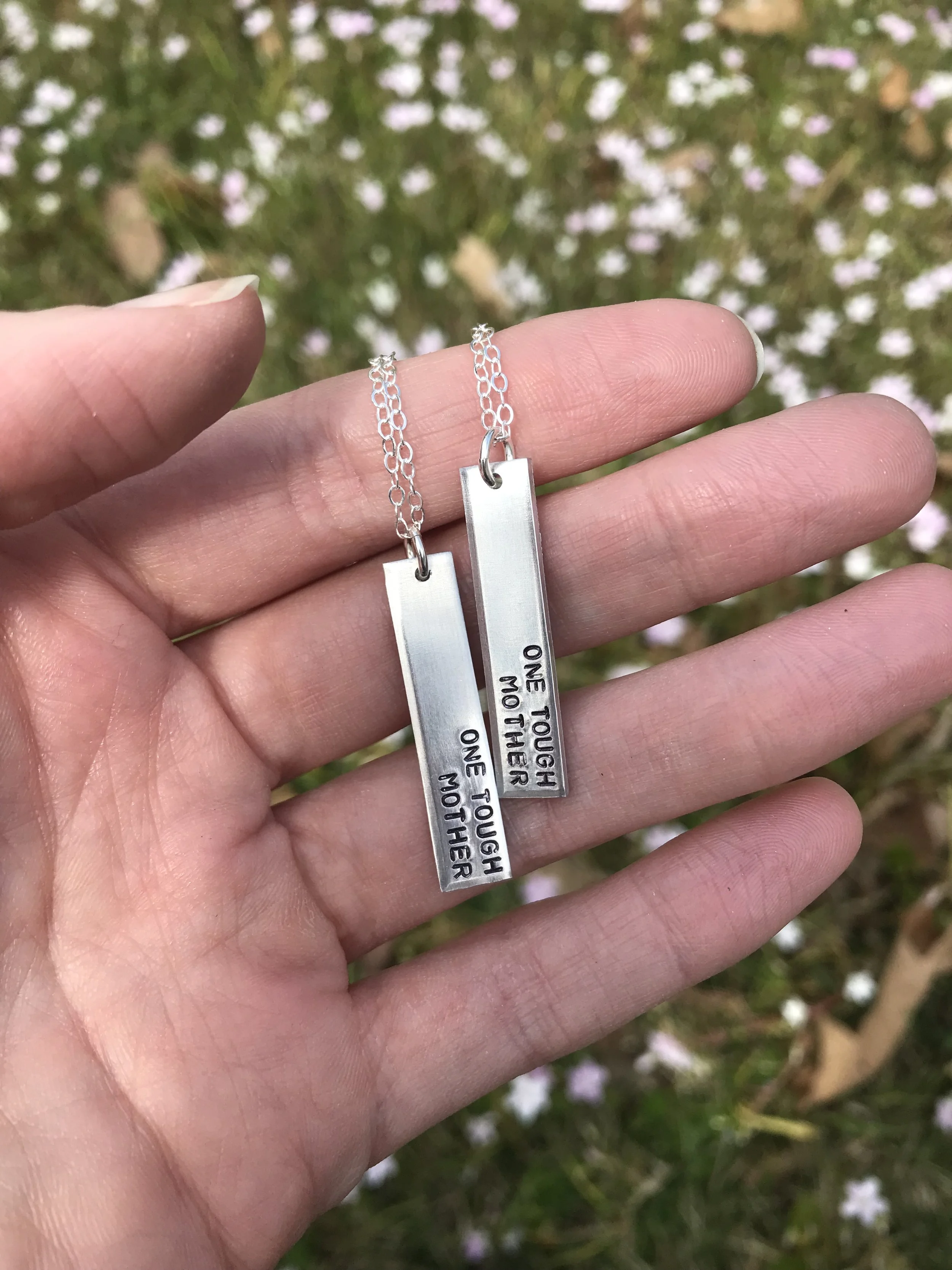 A hand holds out two silver necklaces with silver vertical bar pendants that say "one tough mother"