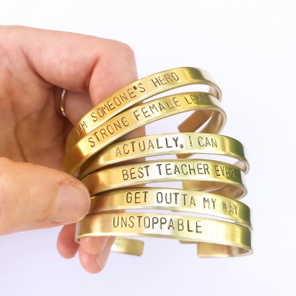 a hand holds out 6 bronze cuffs stamped with different mantras: I'm someone's hero, stong female lead, acutally I can, best teacher ever, get outta my way, unstoppable