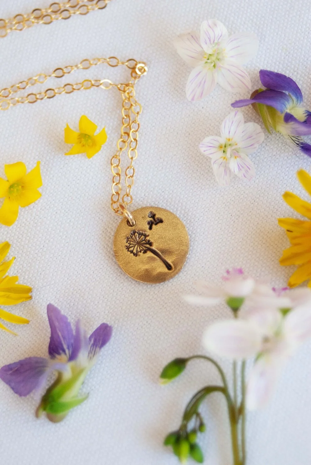 A bronze pendant pressed with the image of a dandelion puff is arranged in wildflowers