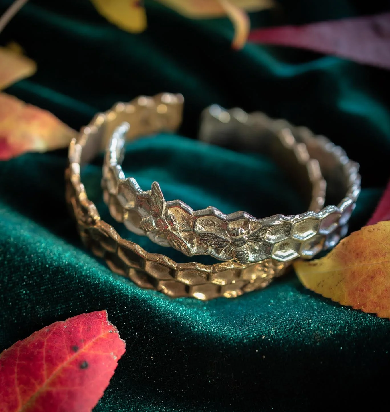 A bronze and a silver cuff stacked on each other over a green fabric. The cuffs are patterned with honeycomb and have bees raised on them.
