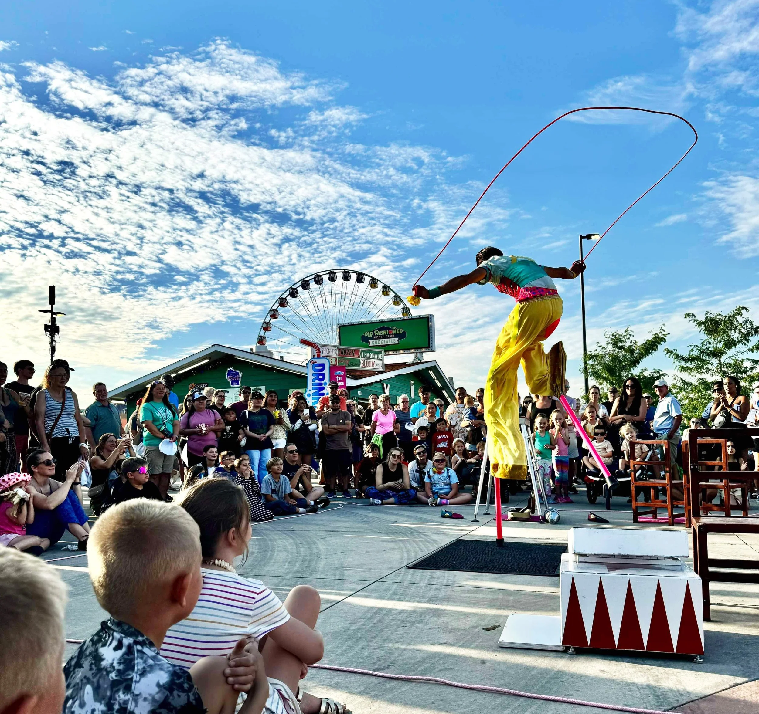 Circus performer on stilts is doing acrobatic stunt at an outdoor State Fair with a crowd of families watching