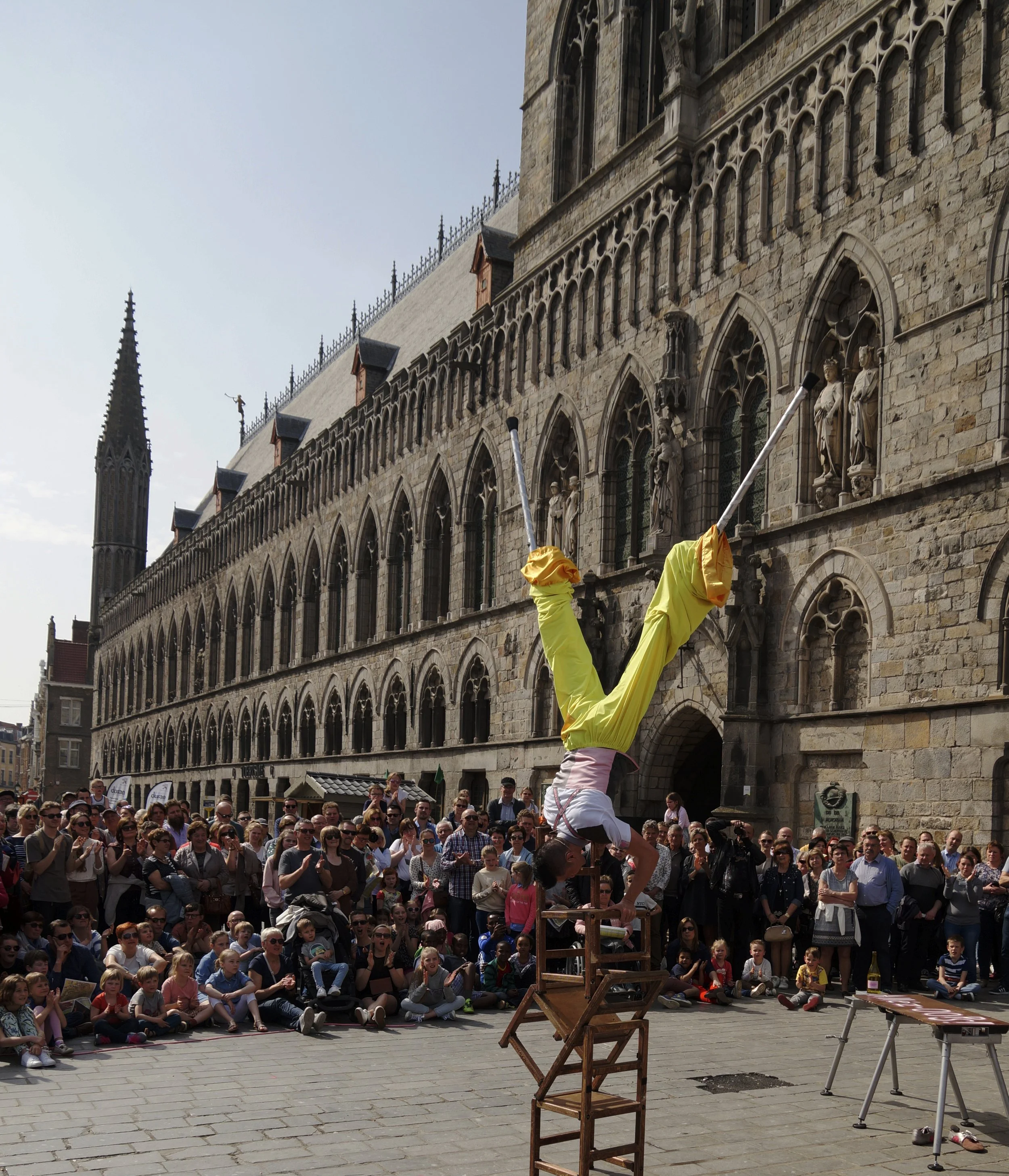 A street performer in yellow pants and white shirt balancing upside-down on their hands on a stack of chairs in front of a Gothic-style building, with a large crowd of spectators watching.