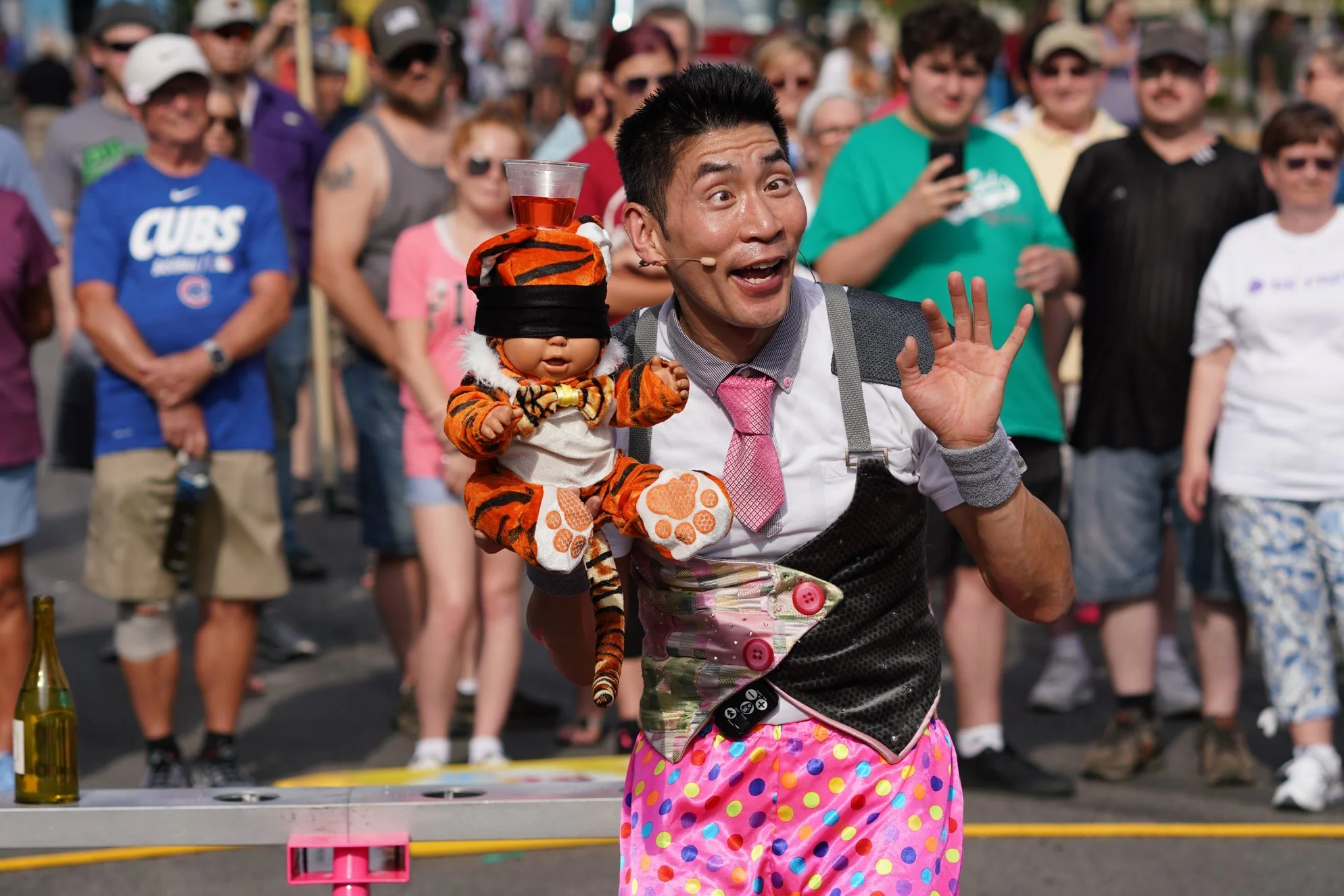 A street performer dressed as a clown with colorful polka dot pants and a pink tie, holding a puppet dressed as a tiger with a drink on its head, in front of a crowd at an outdoor event.