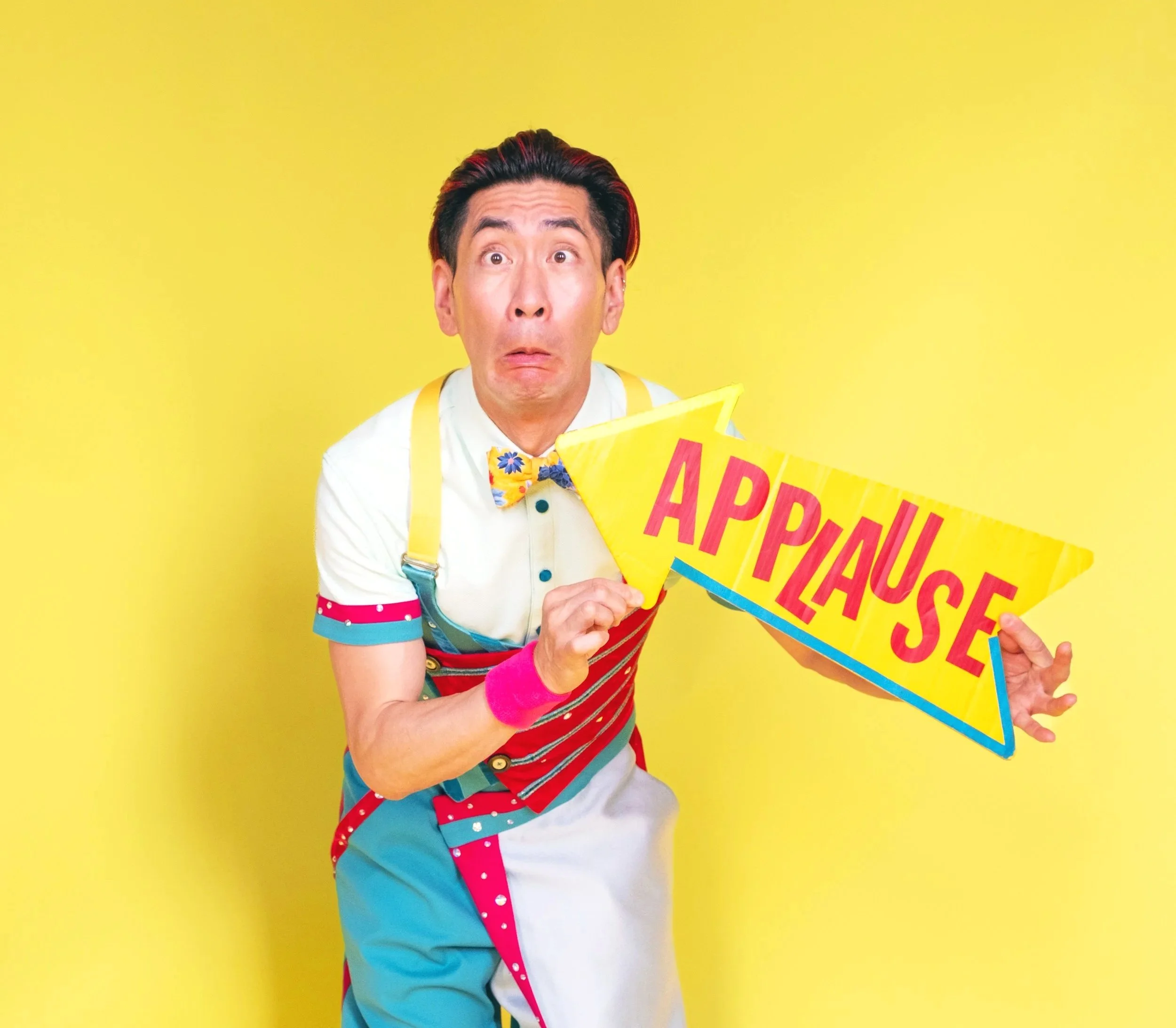 A man dressed as a colorful clown or entertainer holding a yellow sign that says 'APPLAUSE' in a playful font with a surprised or excited facial expression against a bright yellow background.