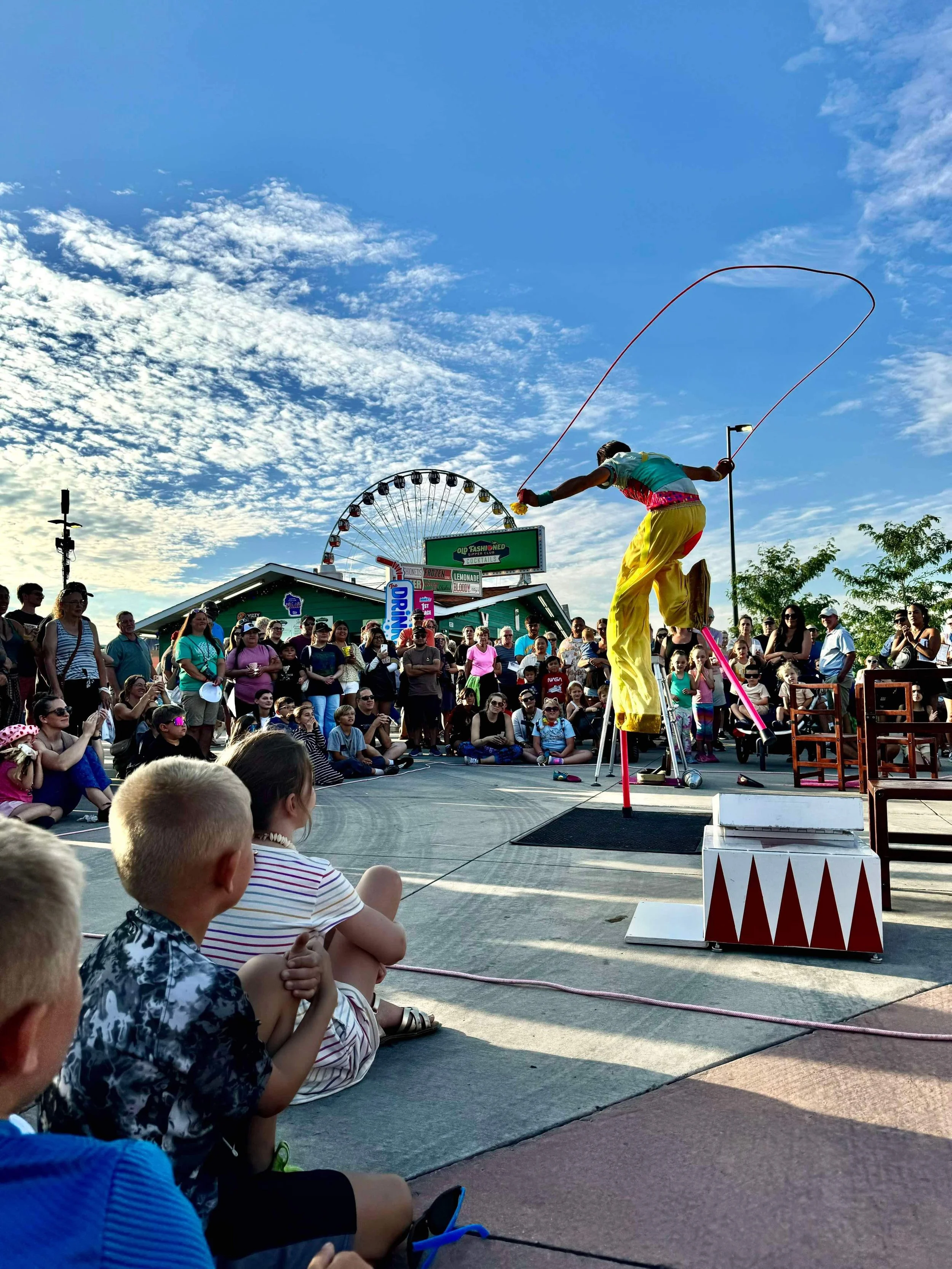 A street performer on stilts performs a jump with a jump rope for a crowd at an outdoor fair, with a Ferris wheel and food stand in the background under a blue sky with clouds.