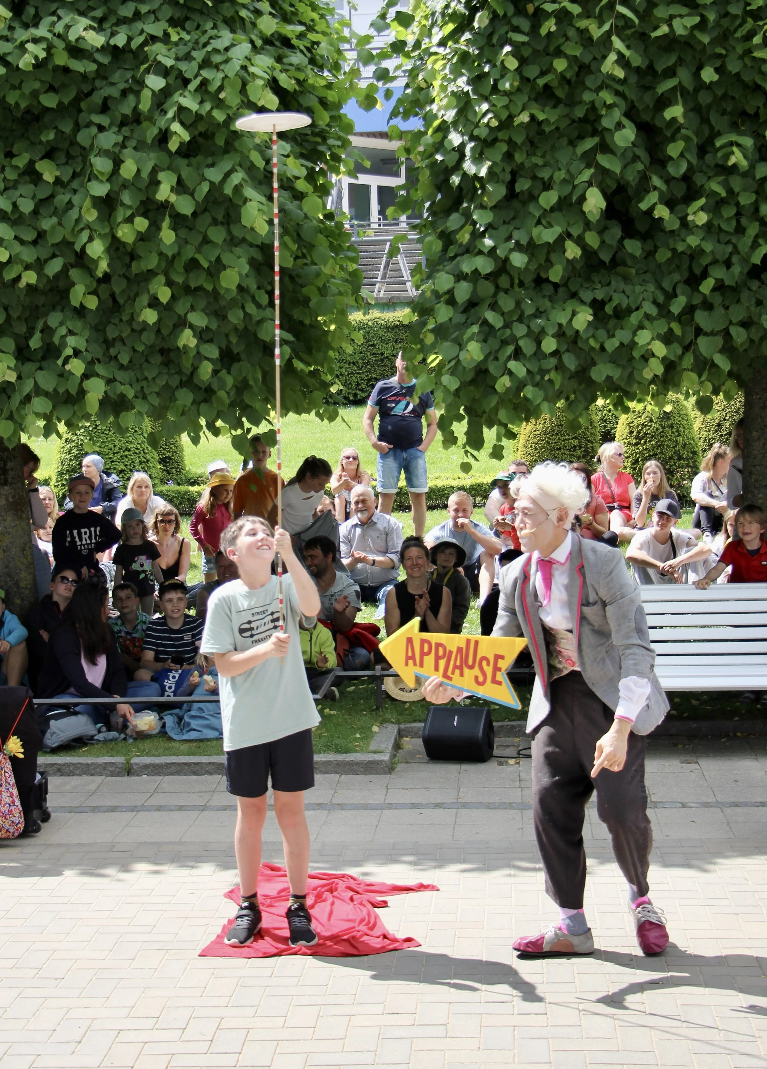 A street performer dressed as an elderly woman with a gray wig, glasses, gray blazer, and dark pants, holding a yellow arrow sign that says 'APPLAUSE'. A young girl is pulling a pole with a red cloth on the ground beneath her. The event is taking place outdoors in front of a crowd sitting on benches and standing, with large trees and a building in the background.
