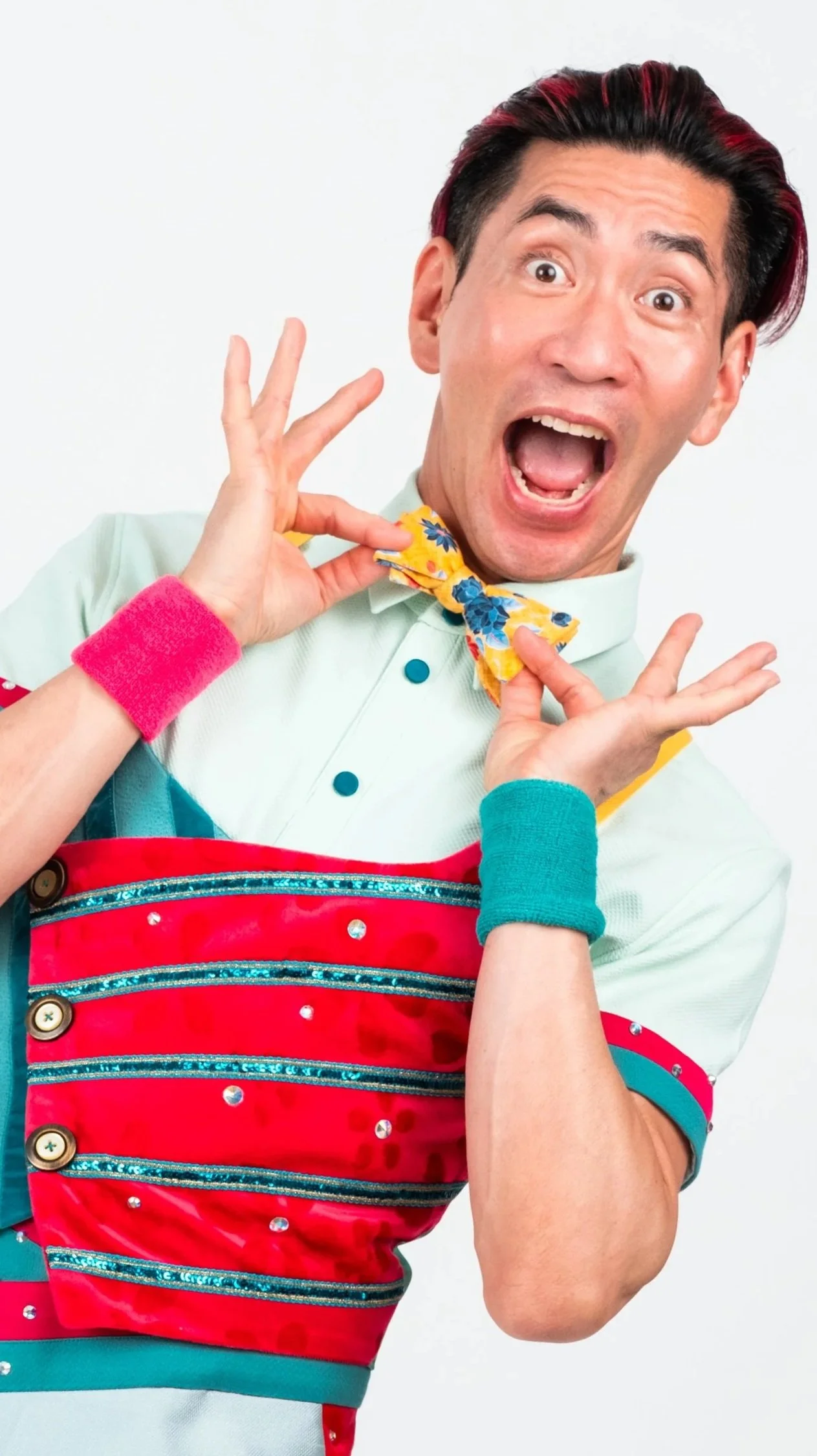 A man with black and red hair wearing colorful clothing, a yellow floral bowtie, and teal and pink accessories, making a surprised or excited facial expression.