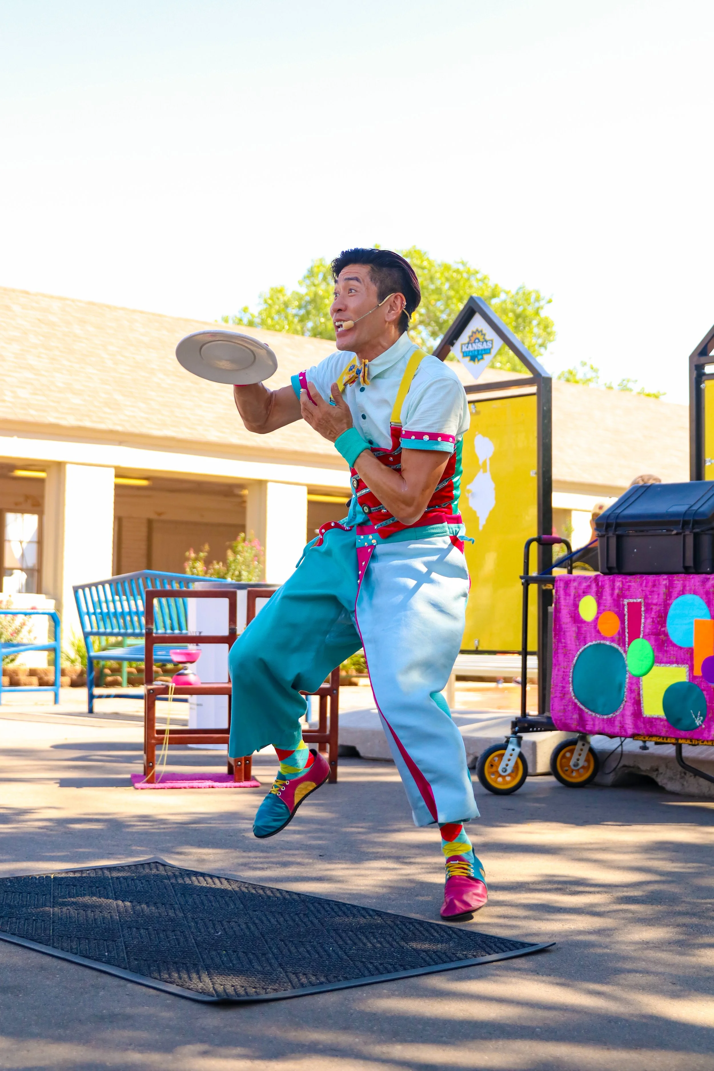 A clown performing a balancing act while standing on one foot, holding a plate, and wearing colorful attire and shoes at an outdoor event.
