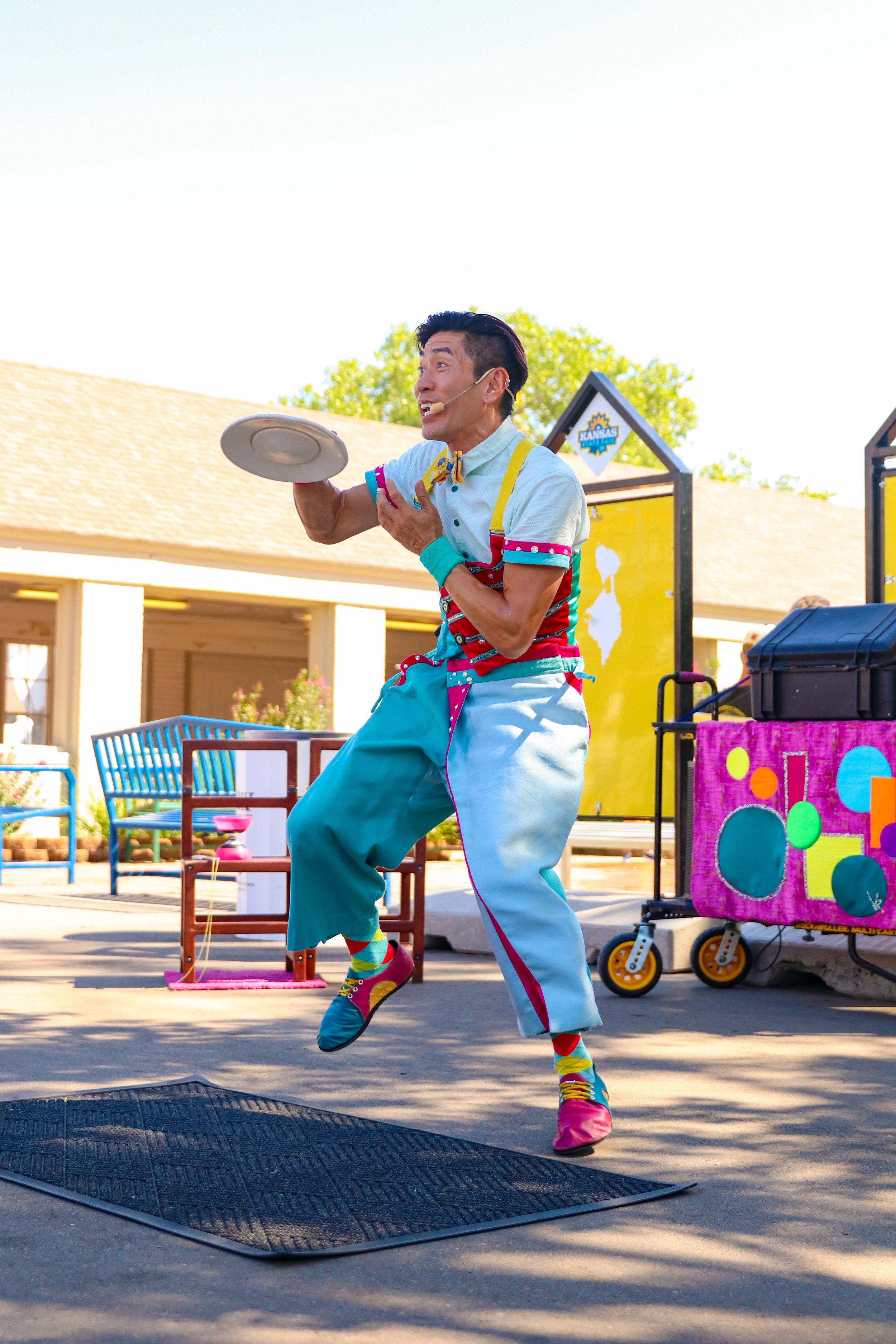 A juggler performing at an outdoor event, wearing colorful clothing and playful shoes, holding a plate, and entertaining the audience.