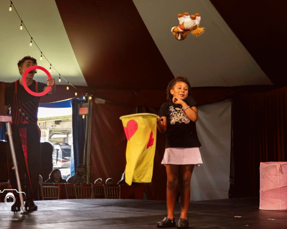 A girl stands on stage during a circus show, with a clown in the background holding a red hoop and a stuffed animal flying from the ceiling.