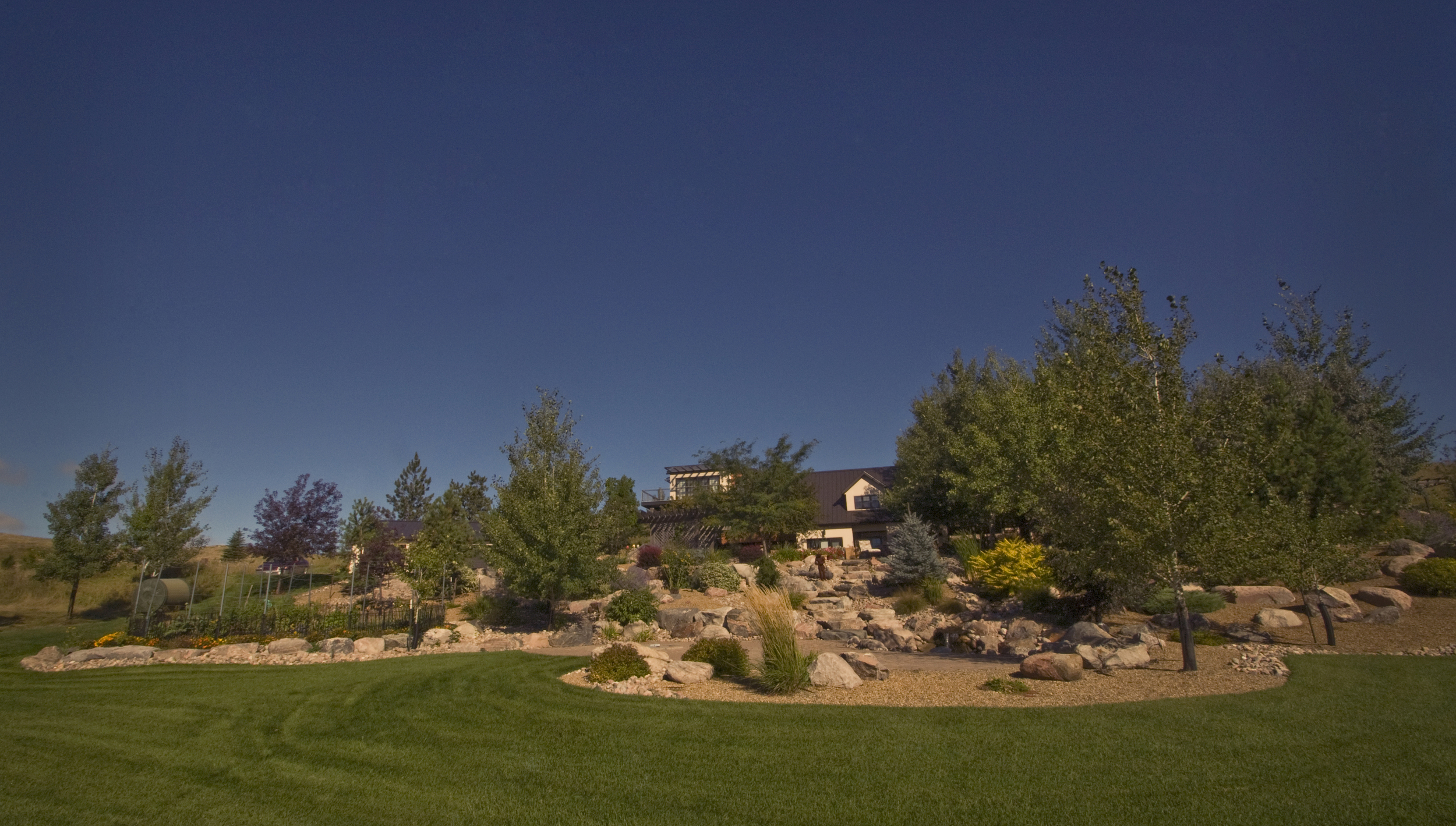  Large rock beds with plantings throughout a backyard. In the center boulder steps cut a path down to a paver patio. 