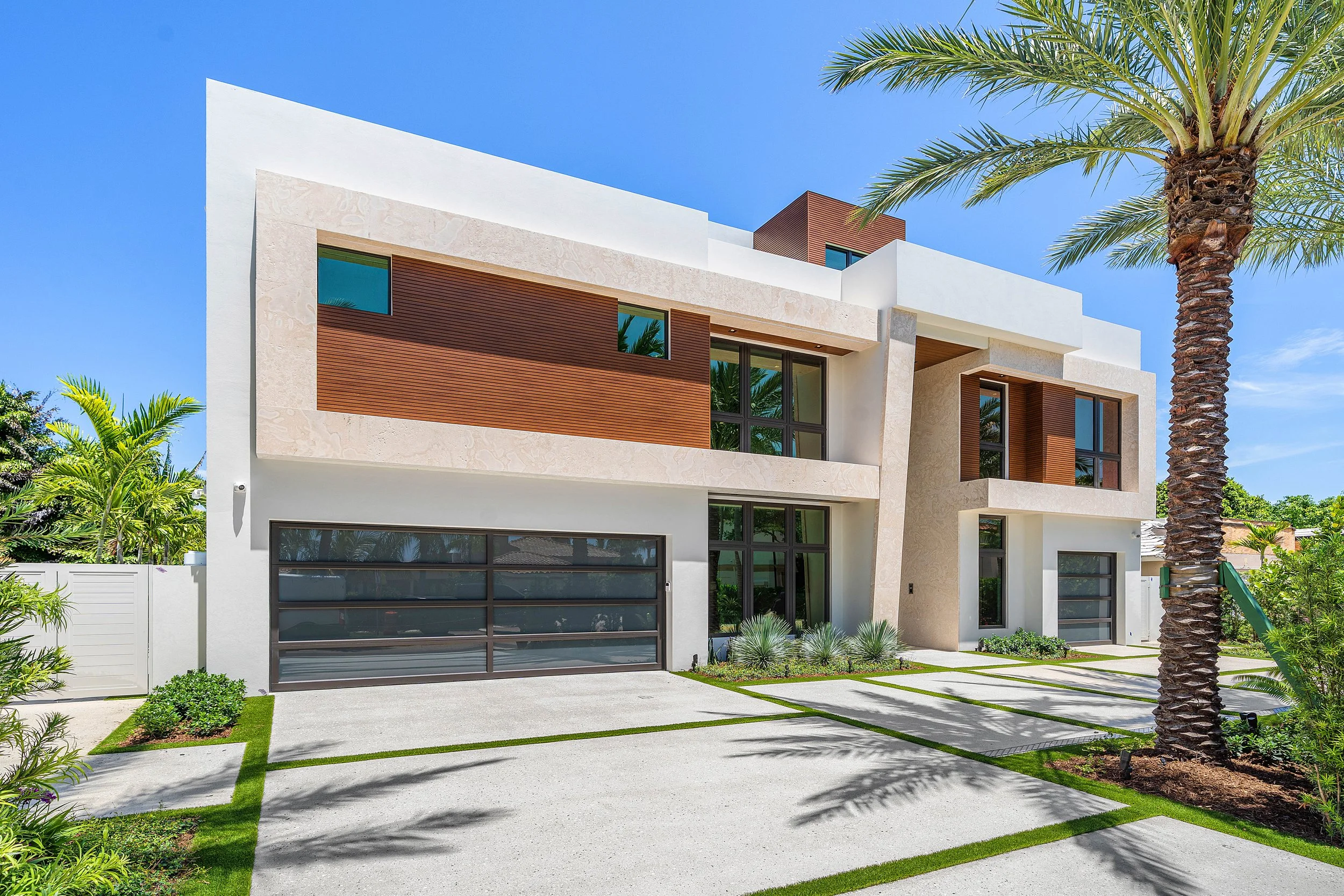 Modern house with white walls, wooden accents, large glass windows, and a garage, surrounded by palm trees and greenery on a sunny day.