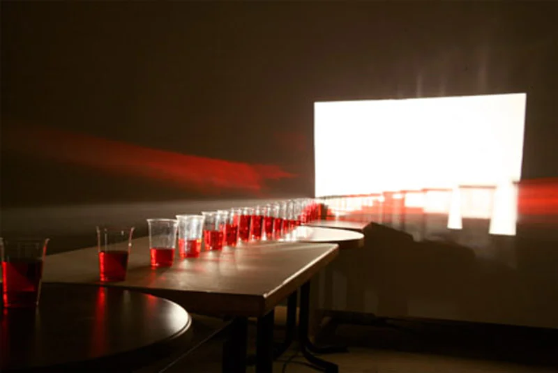 Eulalia Valldosera, El periodo , 2006 Light participatory installation with baby pram. Rails for a cinematographic travelling, trolley’s support with wheels which fit on the rails; slide projector 250W without slide, lenses 70-120mm; baby trolley; 4 tables, 4 chairs, 4 stools; 28 glasses of red wine