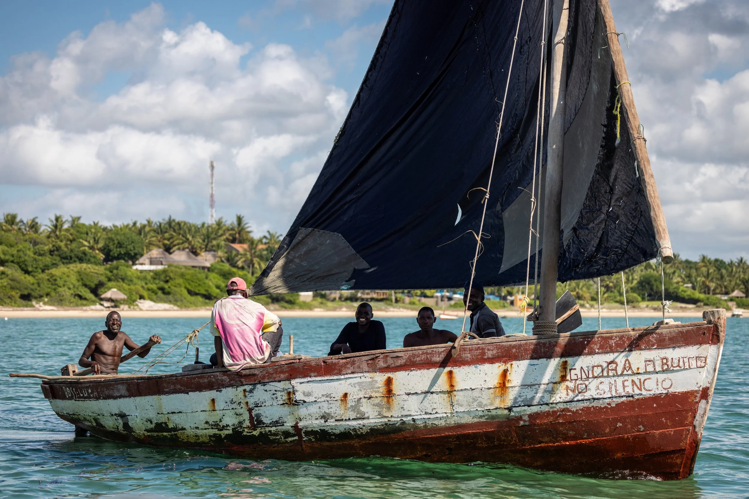 A group of men on a small wooden boat with a black sail, smiling and waving, on a body of water with a lush, green shoreline and cloudy sky in the background.
