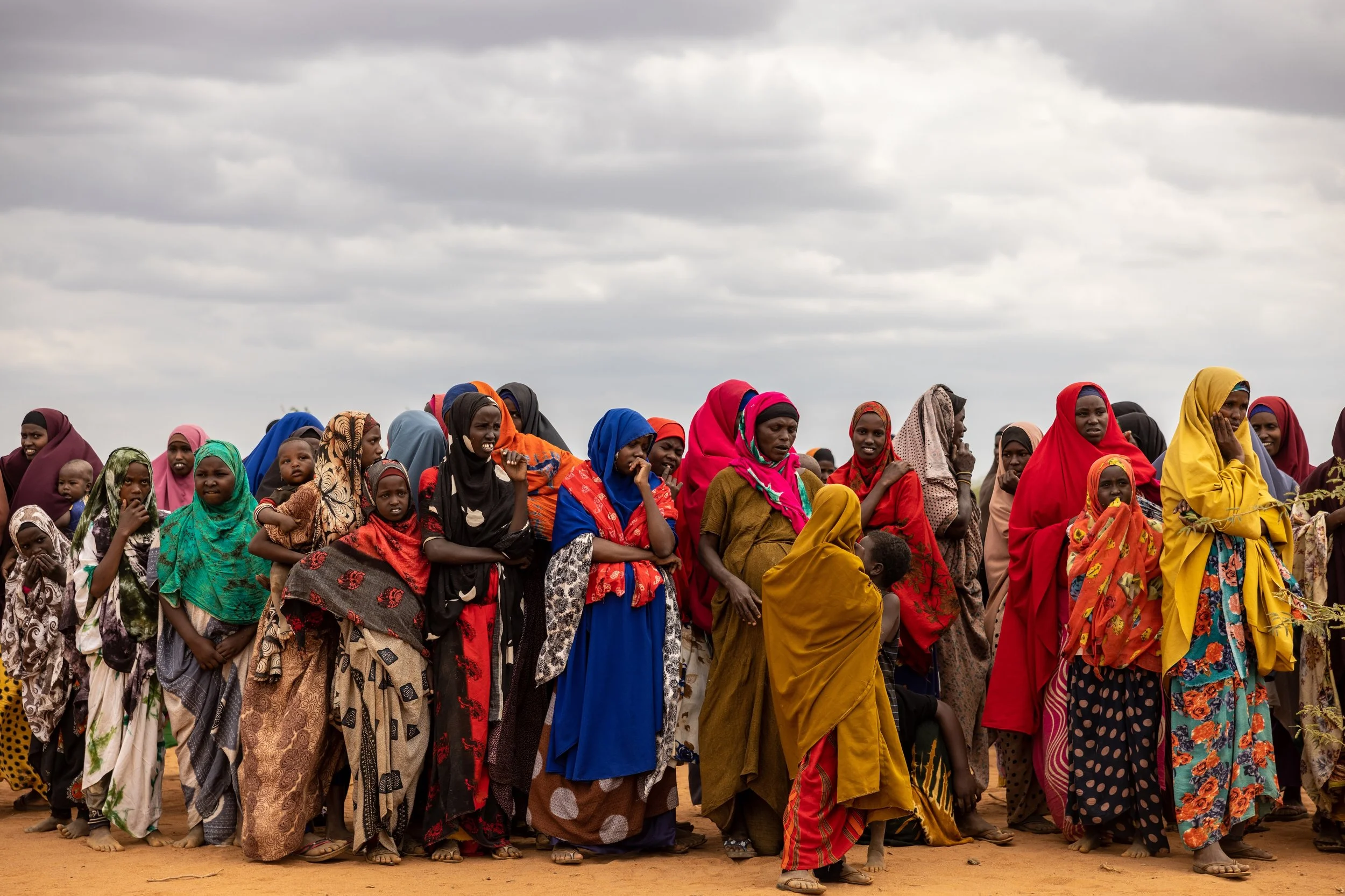 Group of women and children wearing colorful traditional clothing, standing outdoors under cloudy sky.