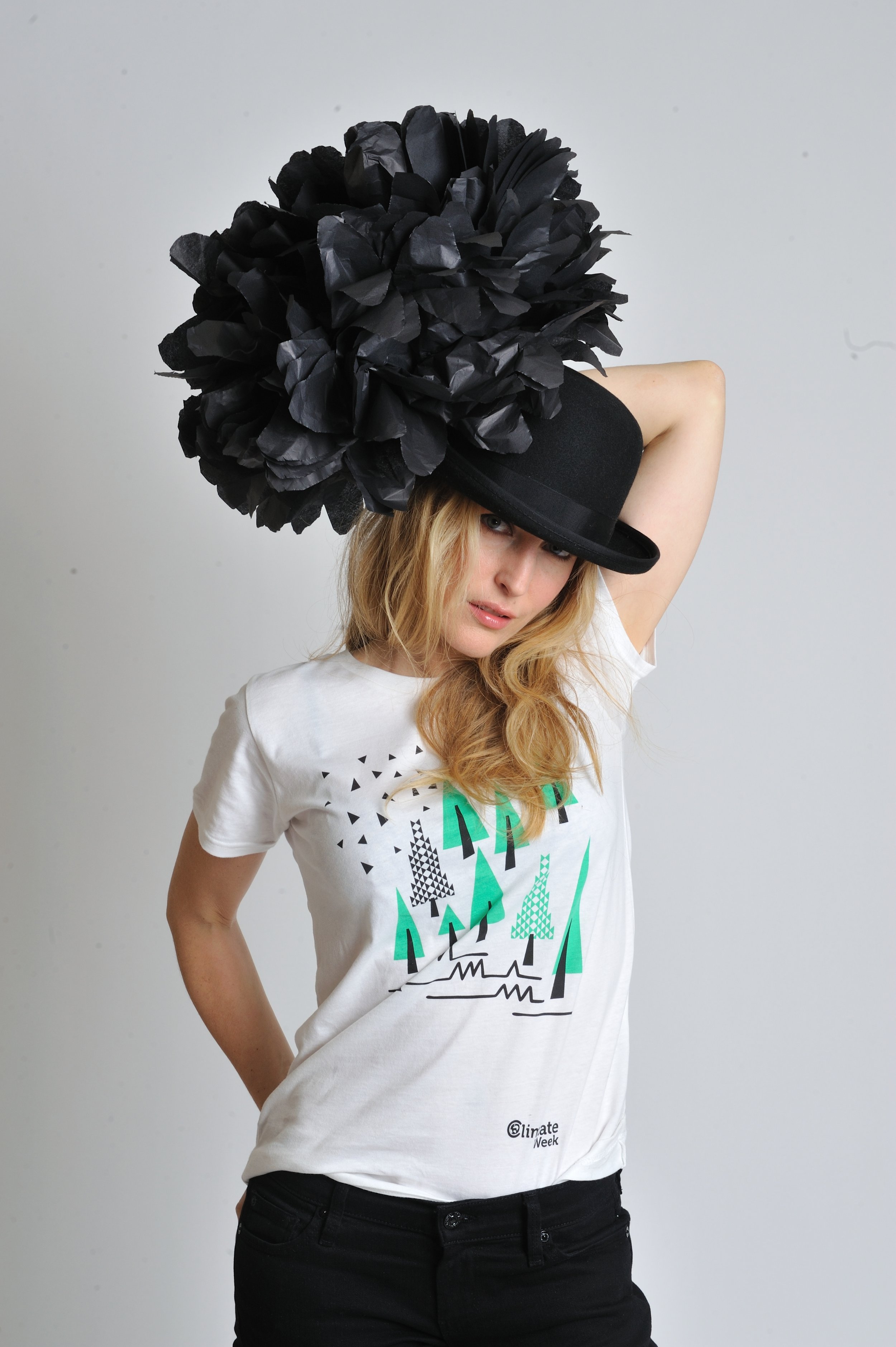A woman with blonde hair is posing in front of a plain light gray background. She is wearing a white T-shirt with colorful graphic design and black pants. She has her right arm raised and resting behind her head, with a large black hat adorned with fluffy black paper flowers on her head.