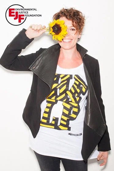 Woman with curly hair smiling and holding a sunflower to her head, wearing a black leather jacket and a white T-shirt with yellow and black lettering.
