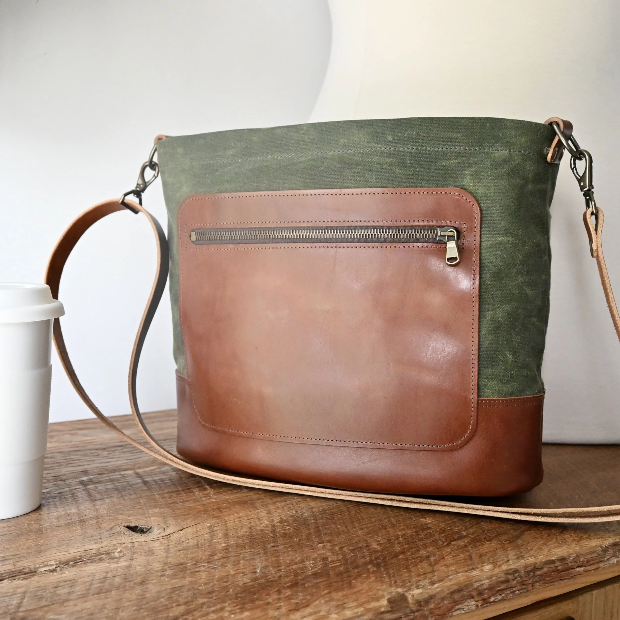 Olive green waxed canvas and warm brown leather Day Bag. The bag is on a wooden table and there is a white coffee cup to the side.