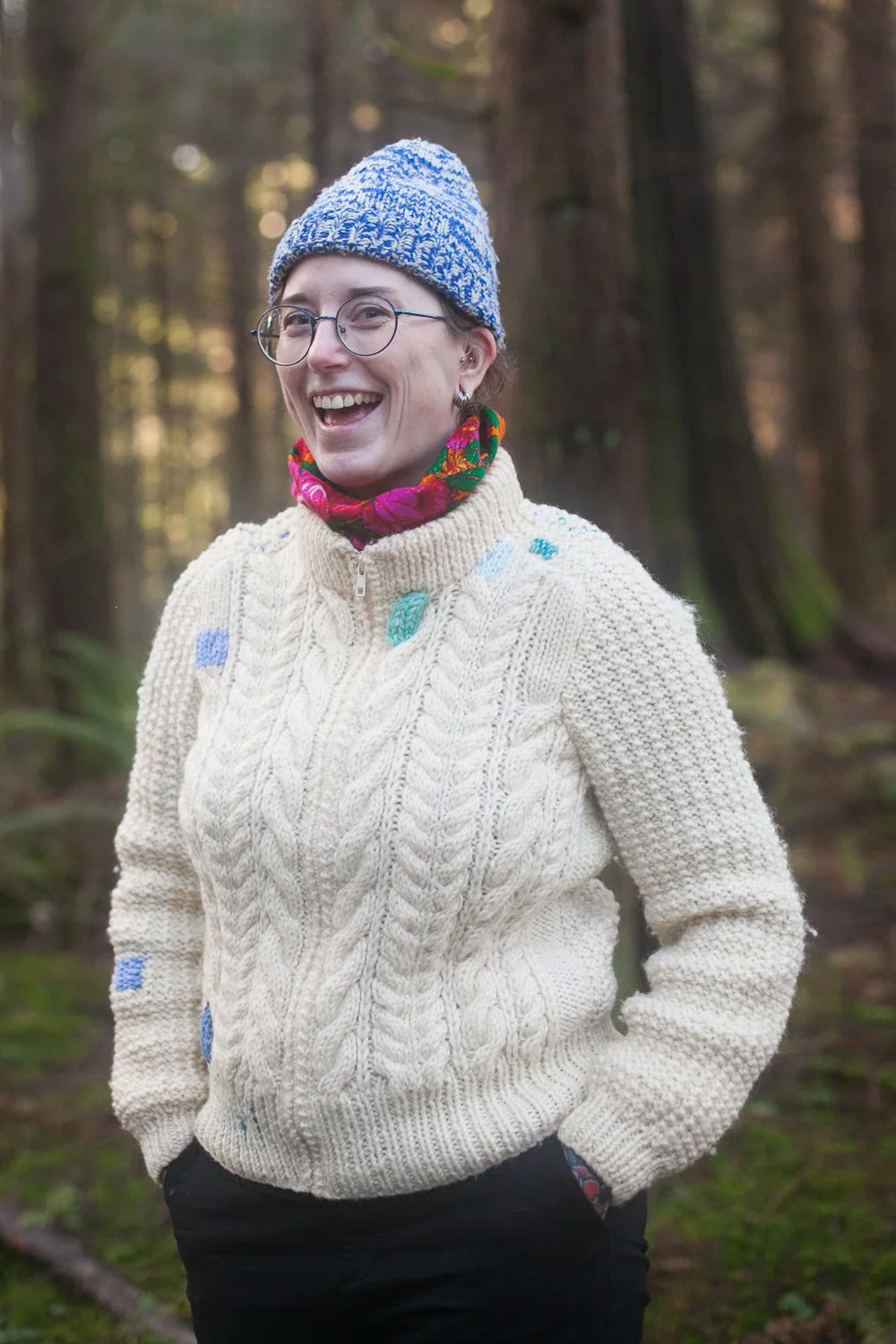 A light-skinned woman is smiling. She has a colourful scarf, a toque, and a white sweater with colourful patches. She's standing in a forest.
