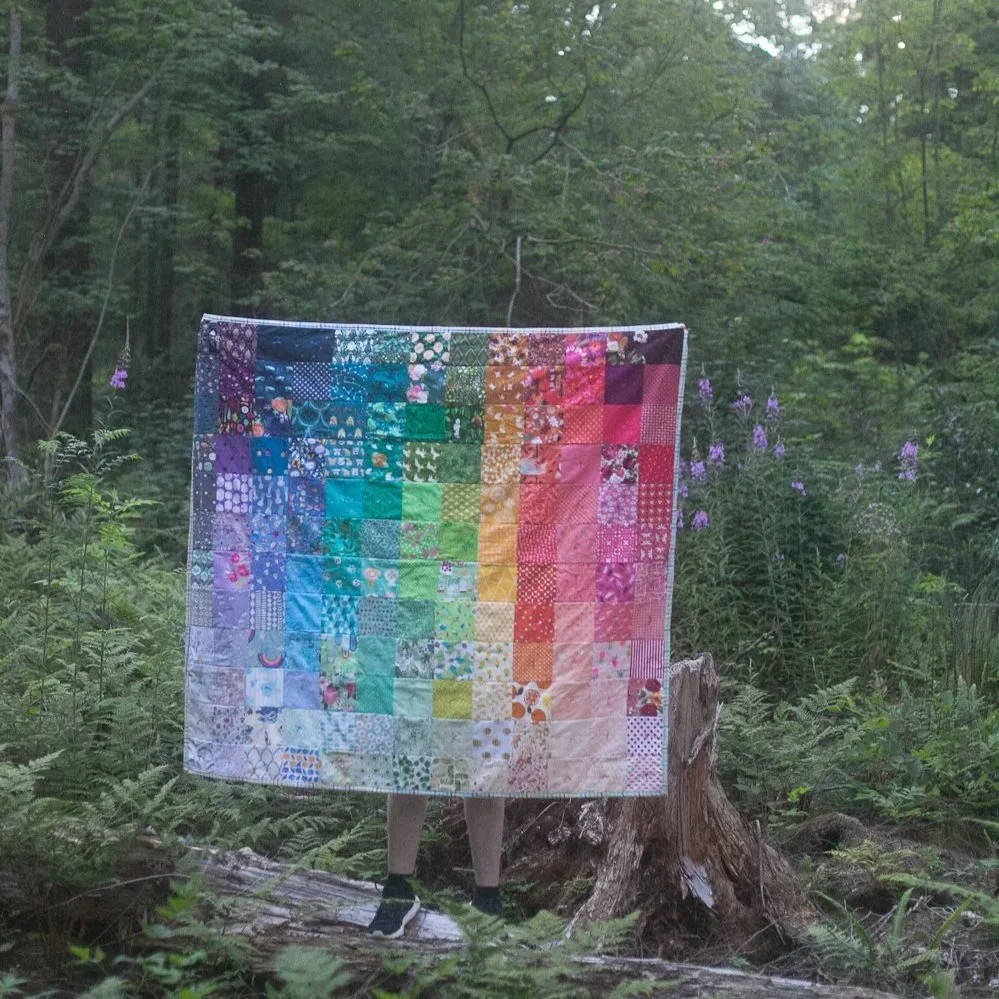 Someone is standing in a forest and holding up a rainbow quilt.