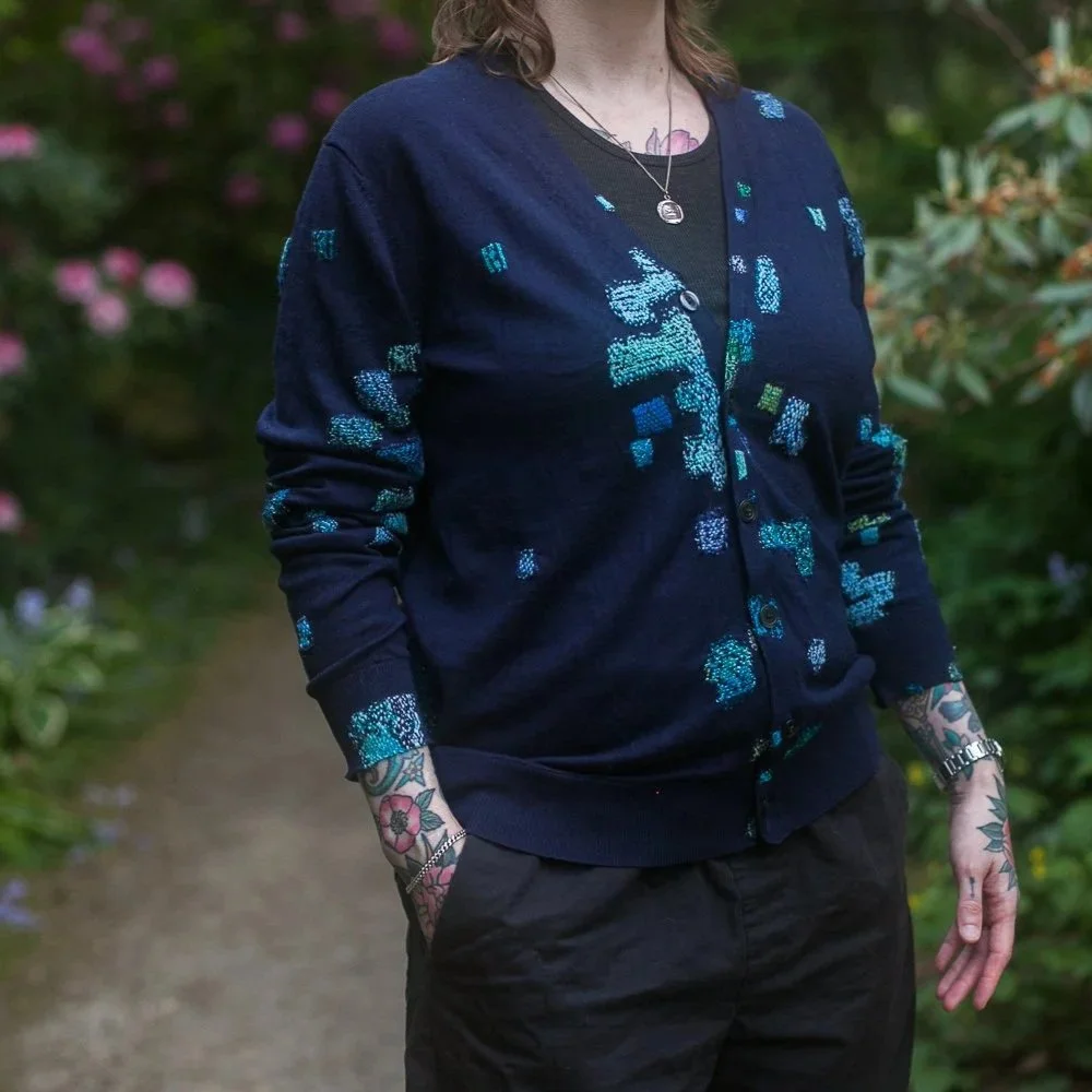 a tattooed woman wearing a blue cardigan that's been heavily mended in shades of blues and greens