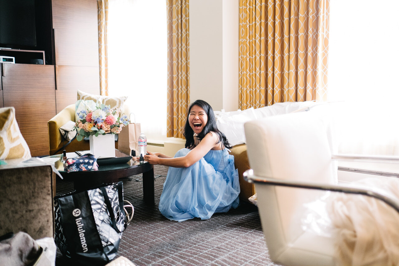  Bridesmaid in blue dress in hotel room laughing while sitting on the floor 