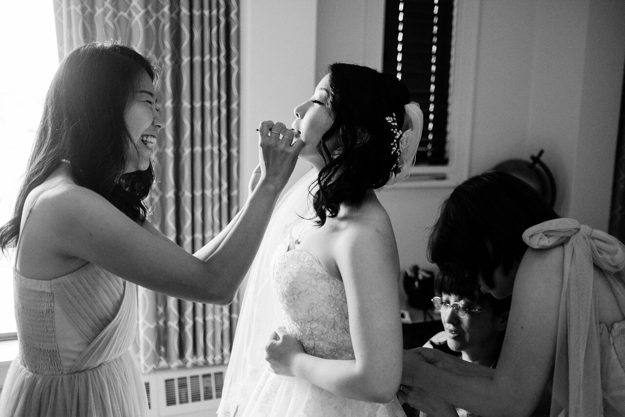  Bridesmaid puts on bride's lipstick while laughing in window light 