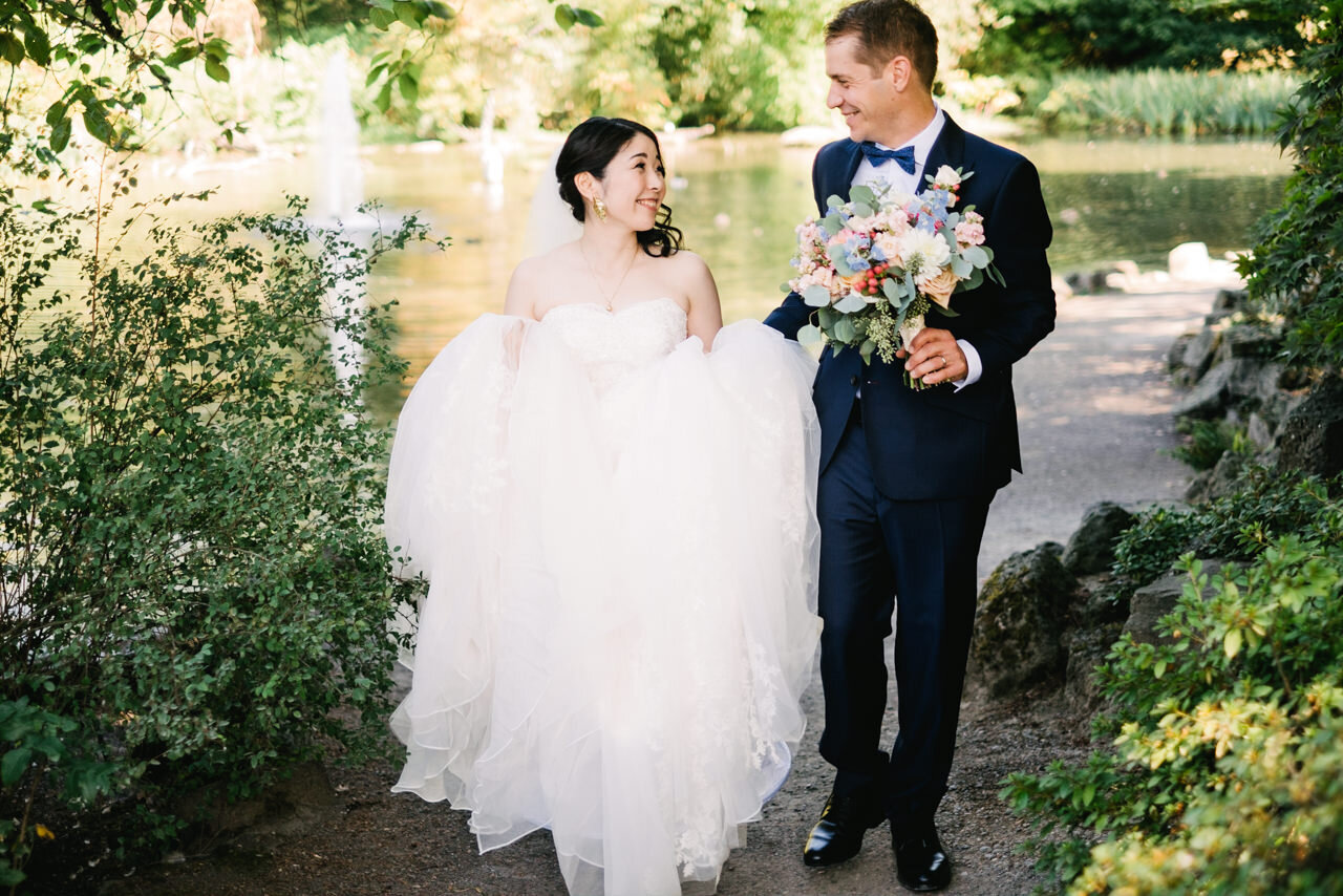  Bride and groom walking together while groom holds peach, blue, and white bouquet with a pond behind them 