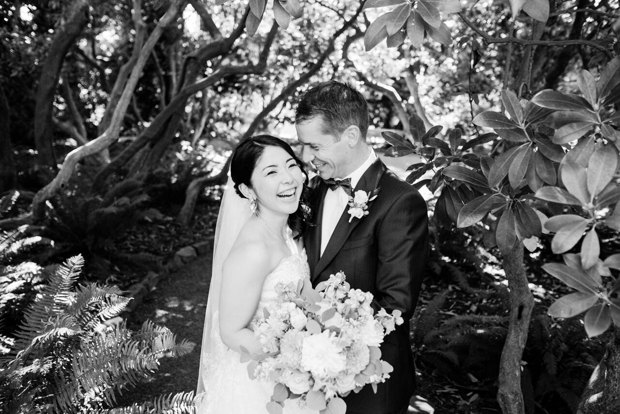  Bride laughs with groom while standing under old rhododendron bushes 