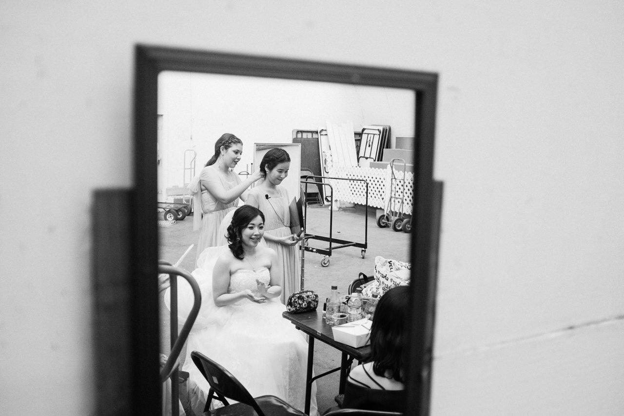  Bride waits for wedding on chair in reflection in mirror while bridesmaids adjust dresses 