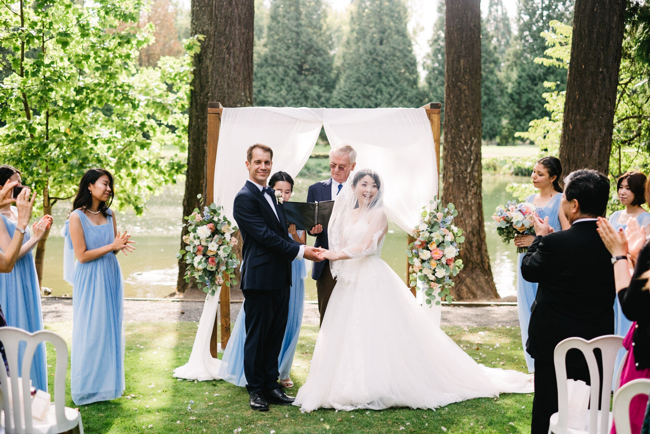  Bride and groom stand in front of wedding arch looking at wedding guests 